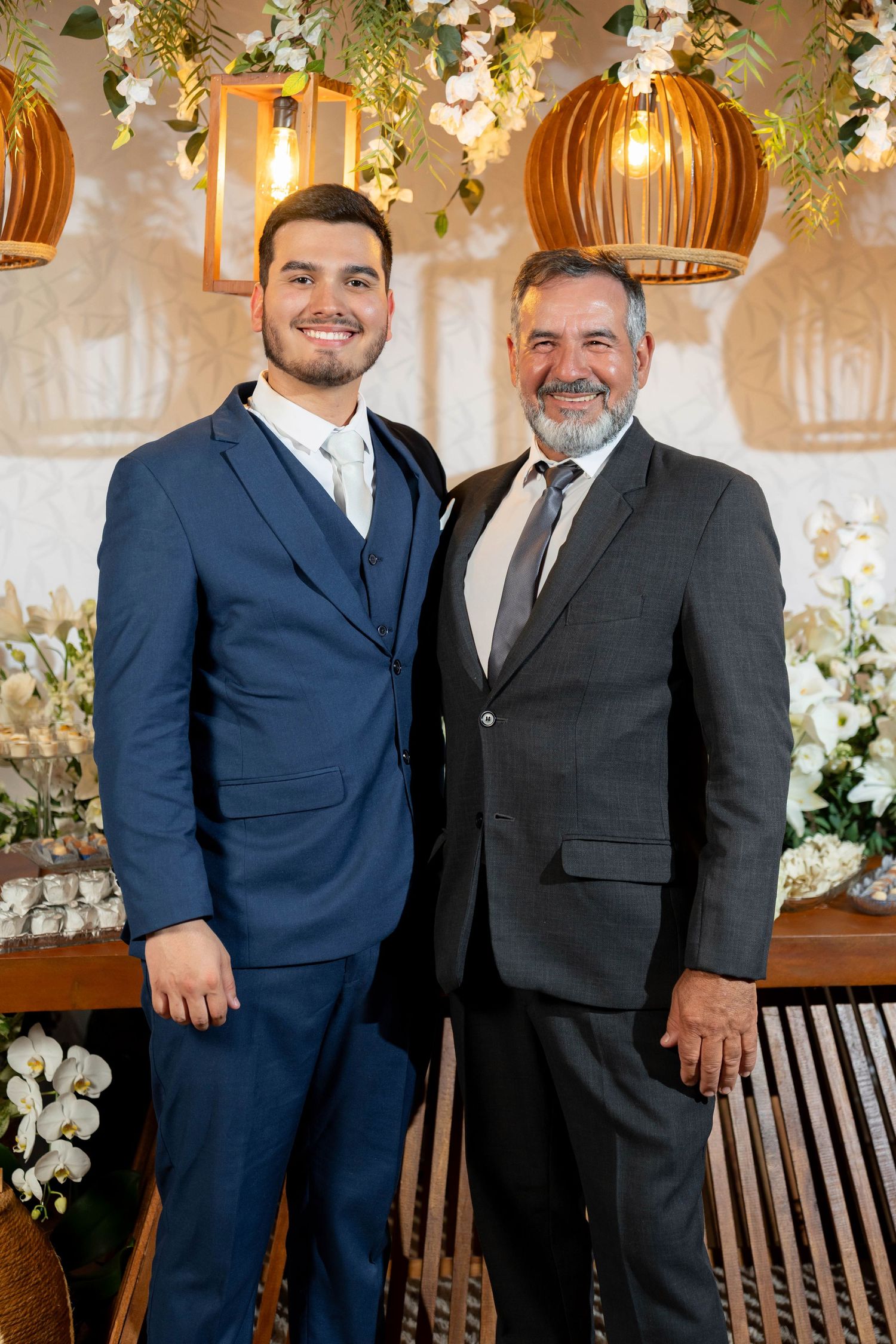 Two men in suits smiling at a formal event with floral decorations and warm pendant lights in the background.