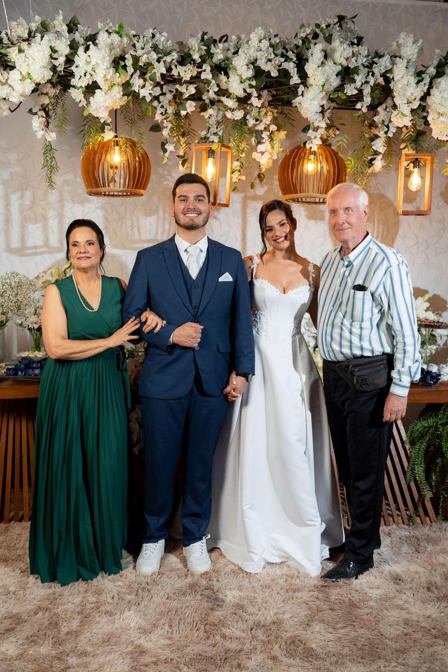 Wedding couple poses with family under floral arch decor, bride in white gown, groom in navy suit, parents in formal attire.
