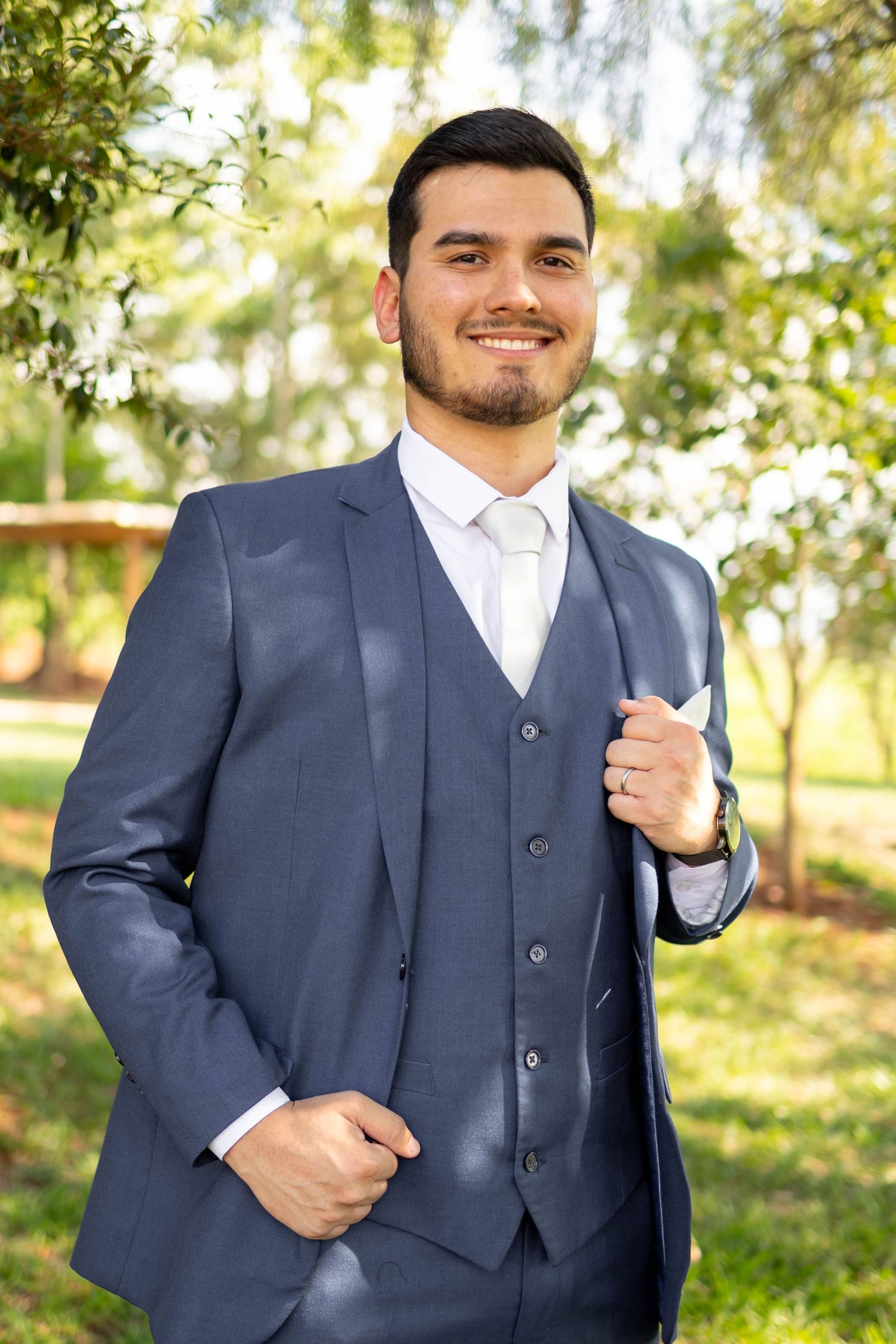 Smiling young man in a navy blue three-piece suit with white tie posing outdoors in a garden setting.