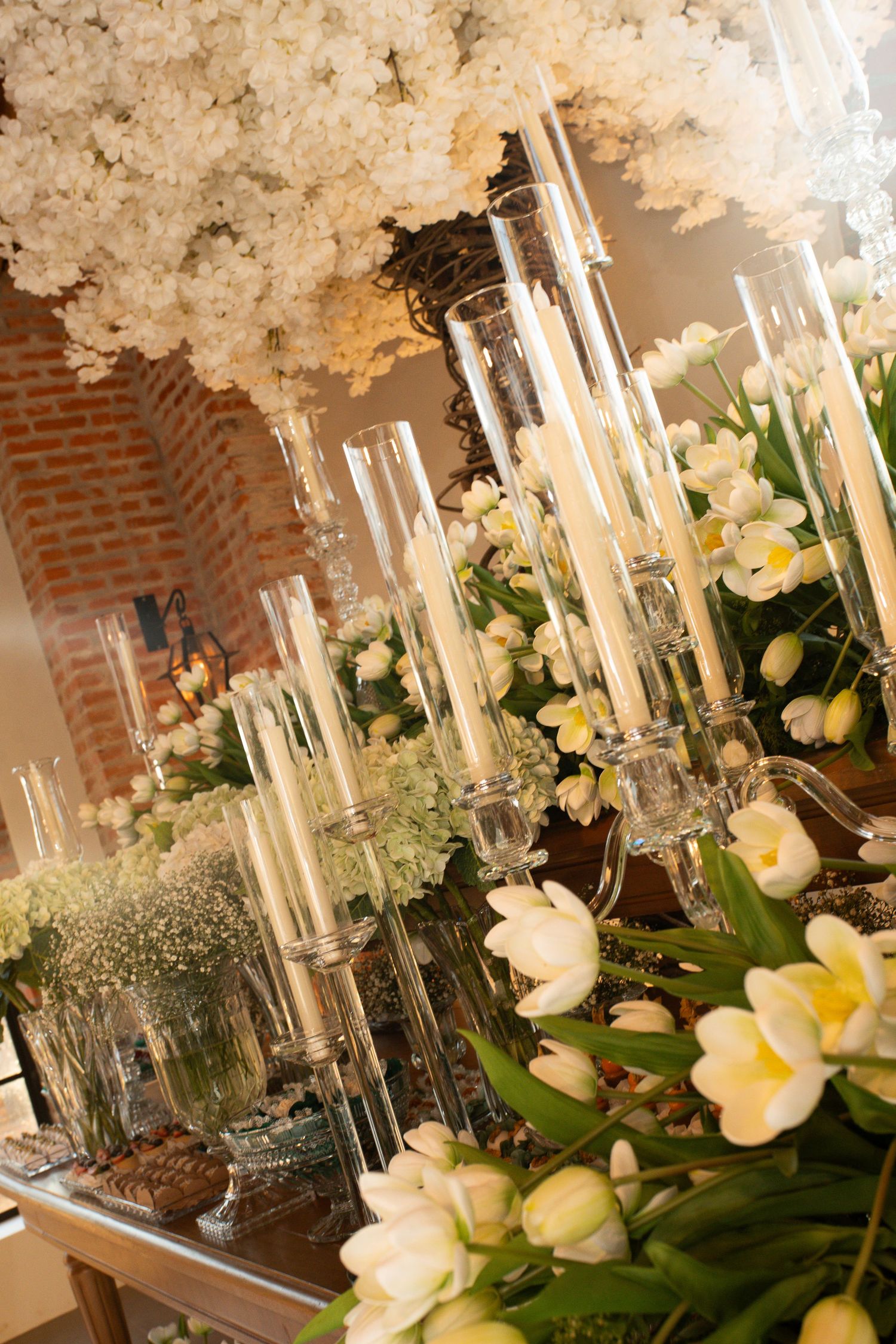 Elegant wedding table with white tulips, tall glass candle holders, and lush floral arrangements in a warmly lit venue.