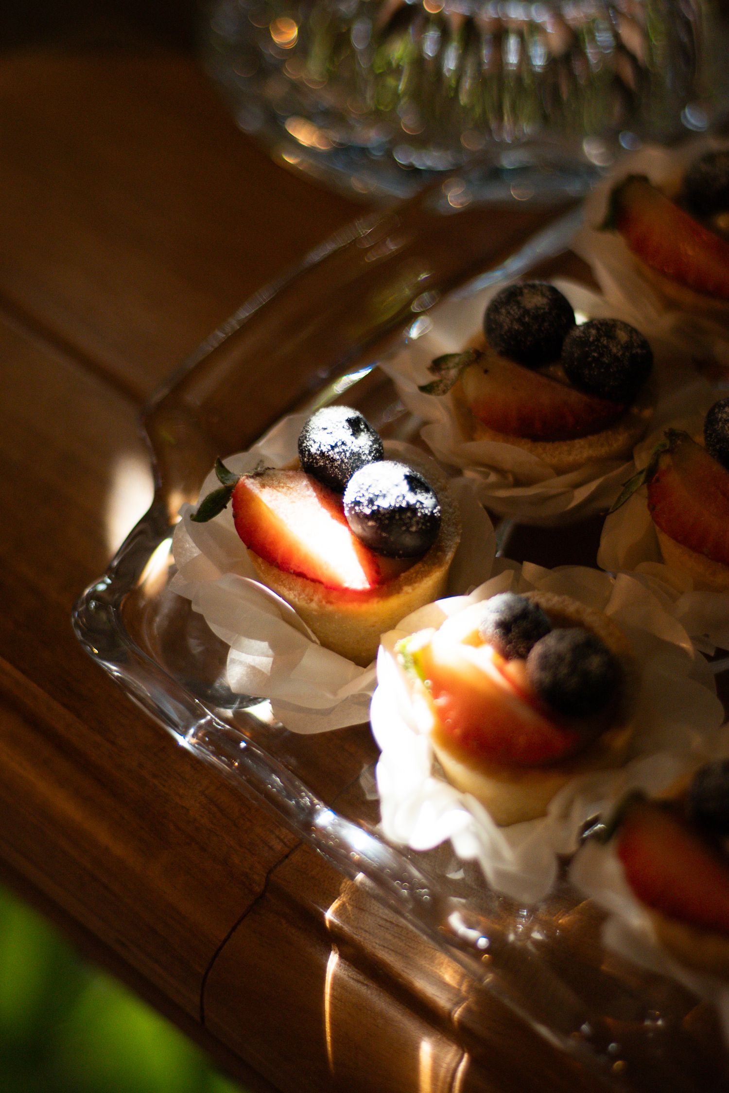 Elegant mini fruit tarts topped with blueberries and peach slices on a glass serving tray, dusted with powdered sugar.