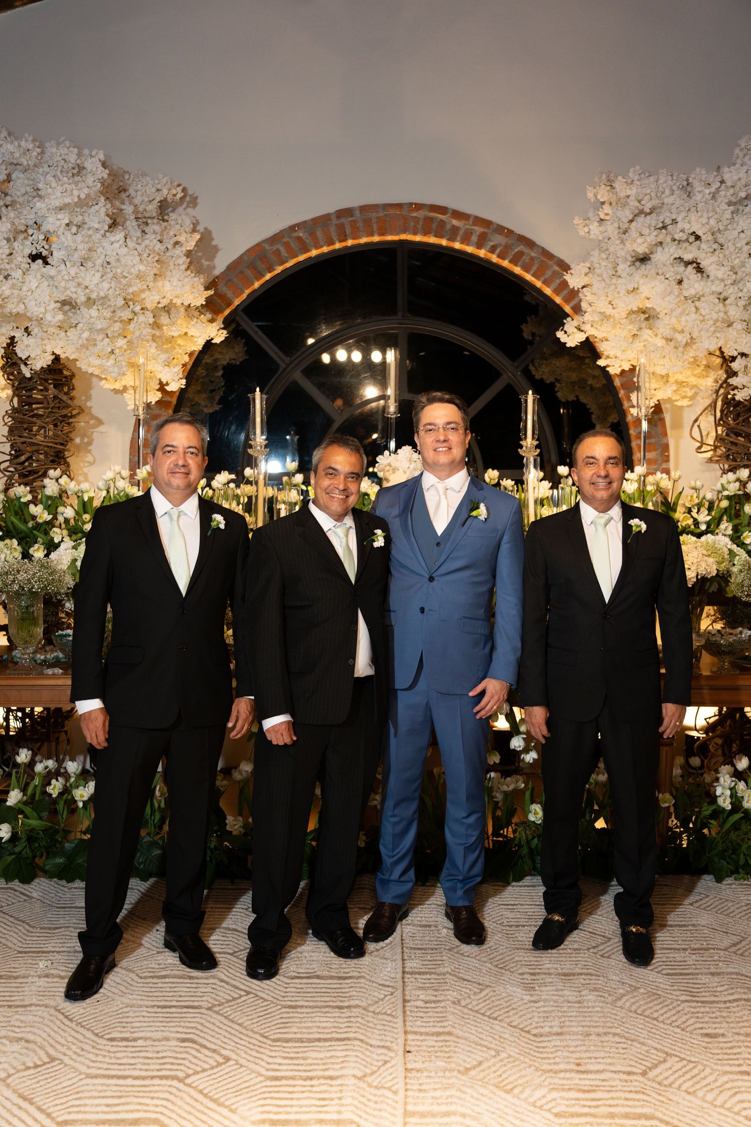Four men in formal suits pose together at an elegant wedding venue adorned with white floral arrangements.