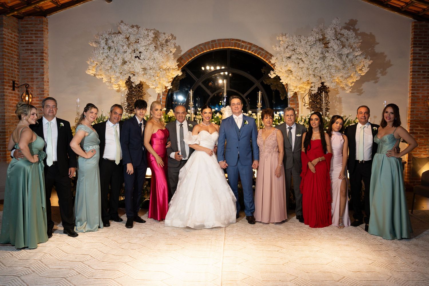 Wedding party poses together in elegant venue with floral arch, bride in white gown surrounded by guests in formal attire.