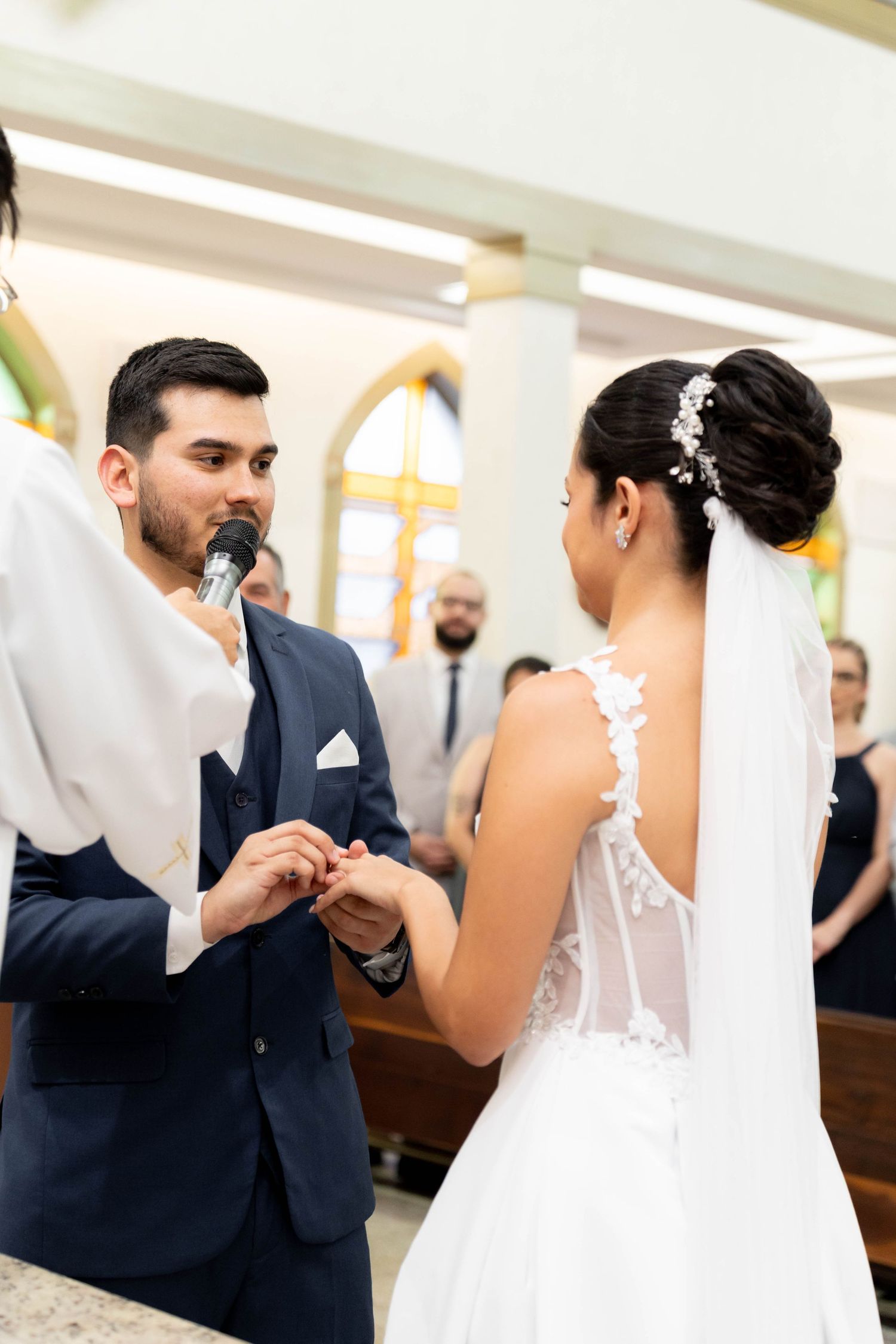 Groom placing wedding ring on bride's finger during church ceremony, both dressed in formal wedding attire.