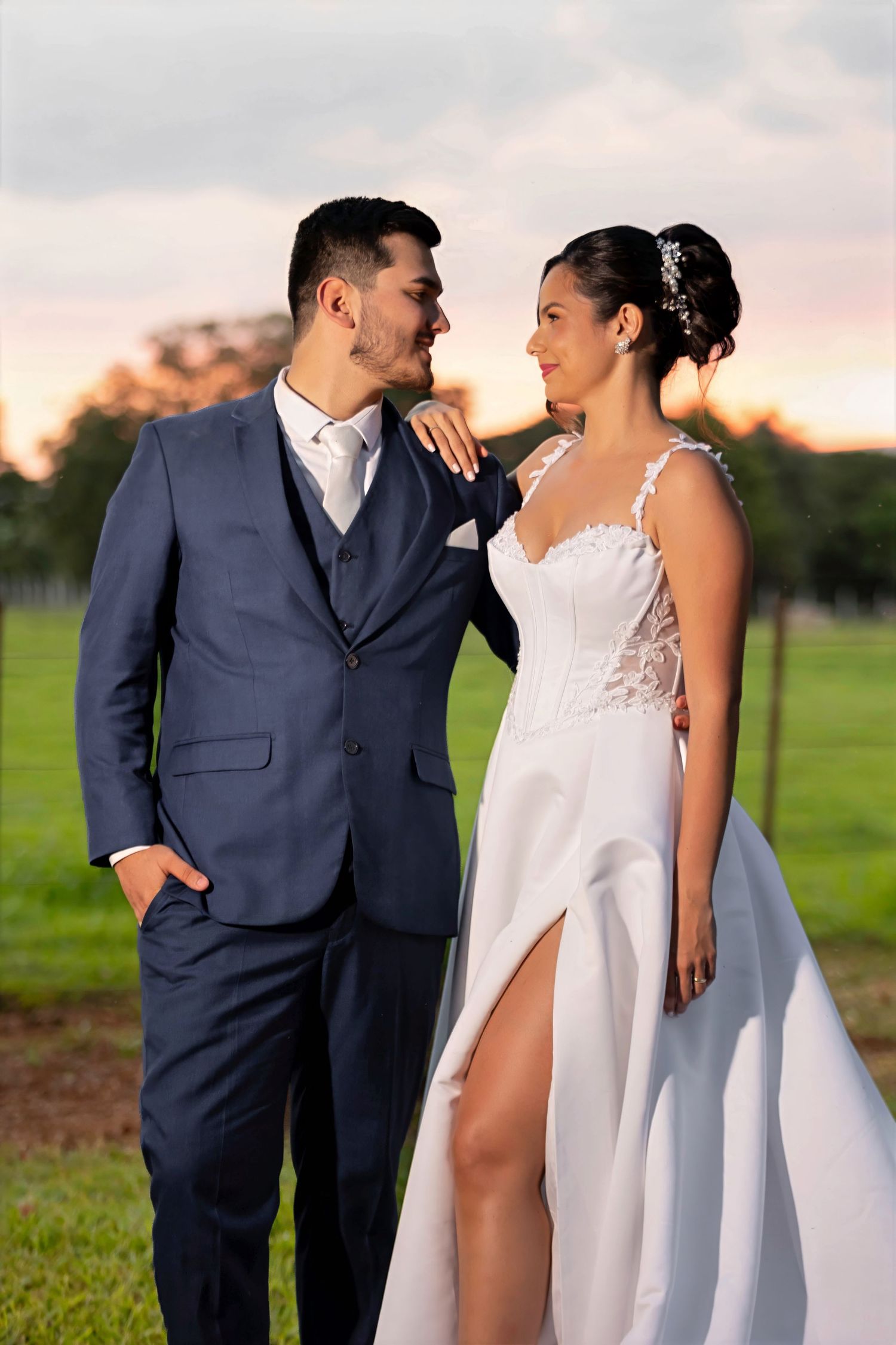 Bride in white lace gown and groom in navy suit share a romantic gaze at sunset on a green lawn.