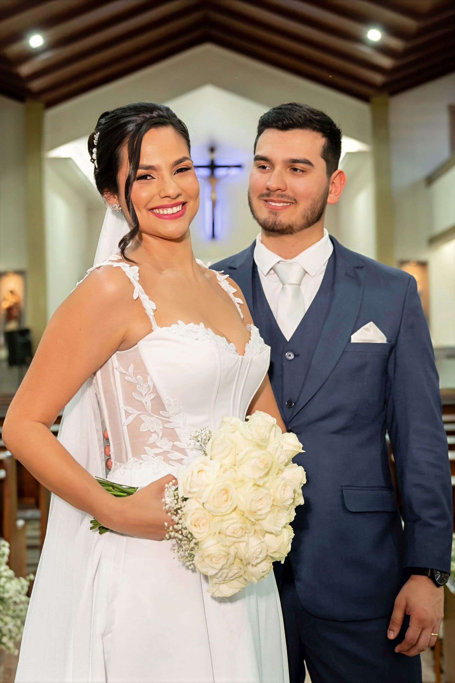 A smiling bride in a white gown holds white roses beside a groom in a navy suit inside a church.