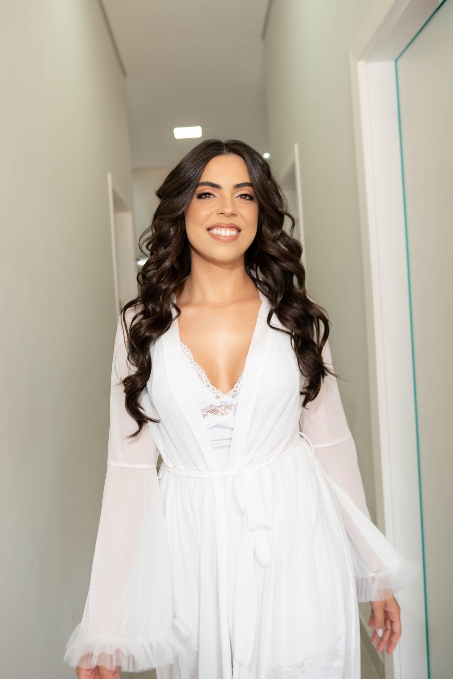Smiling woman with long dark wavy hair wearing a white lace bridal robe standing in a bright hallway.