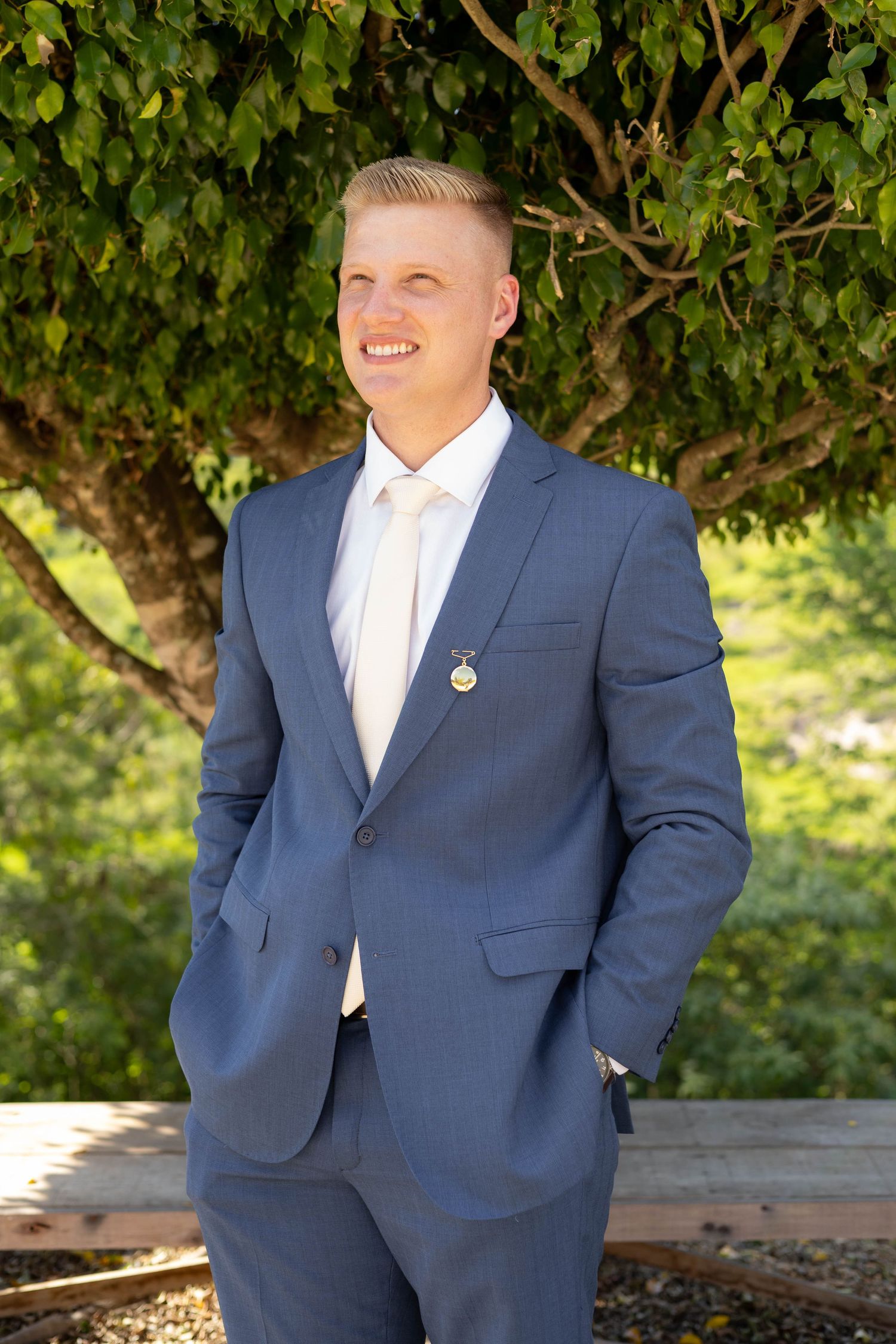 Young man in blue suit and white tie smiling outdoors near green trees.