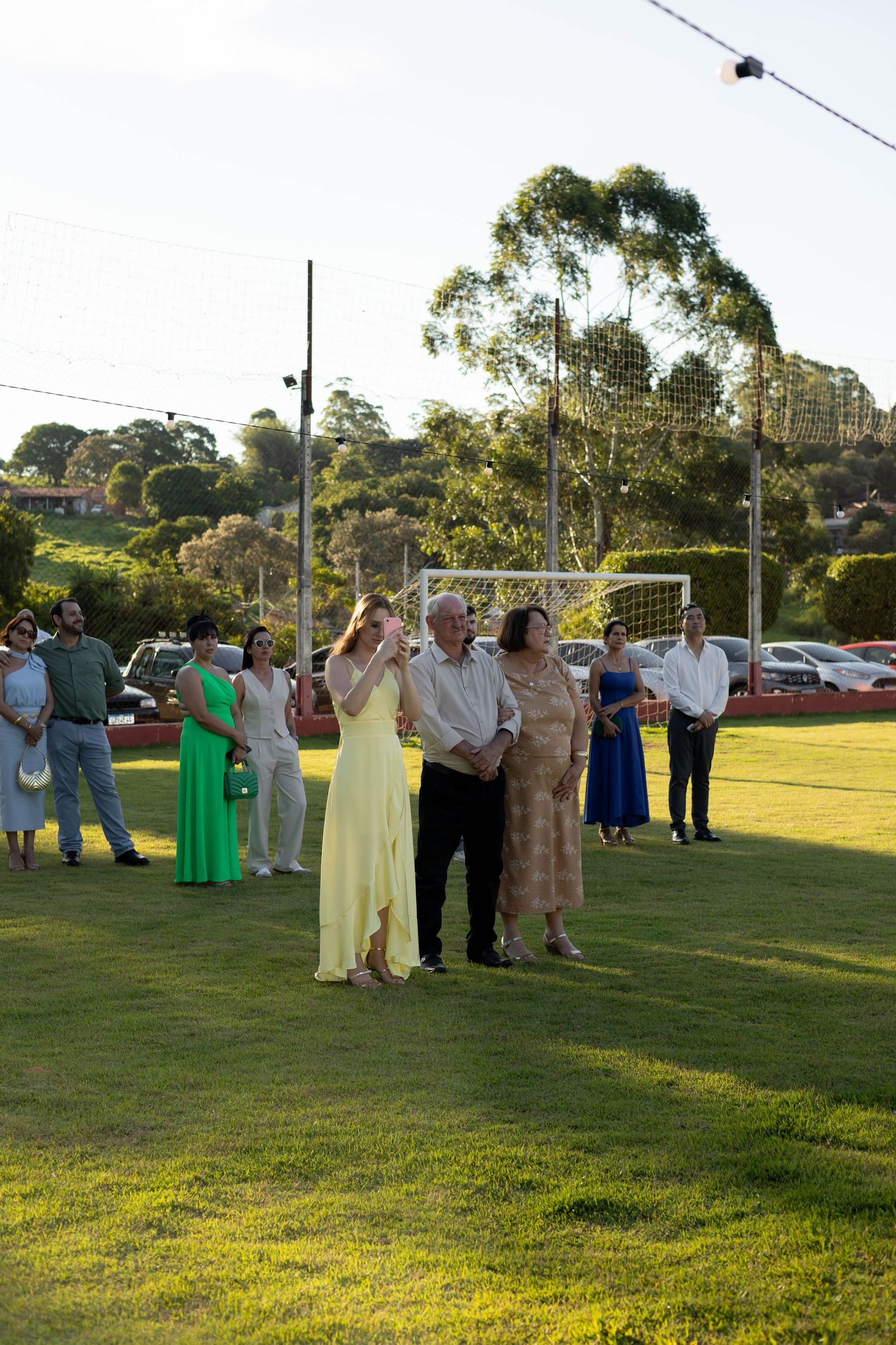 Guests in formal attire gather on a green lawn at an outdoor wedding reception with trees and a soccer goal in the background.