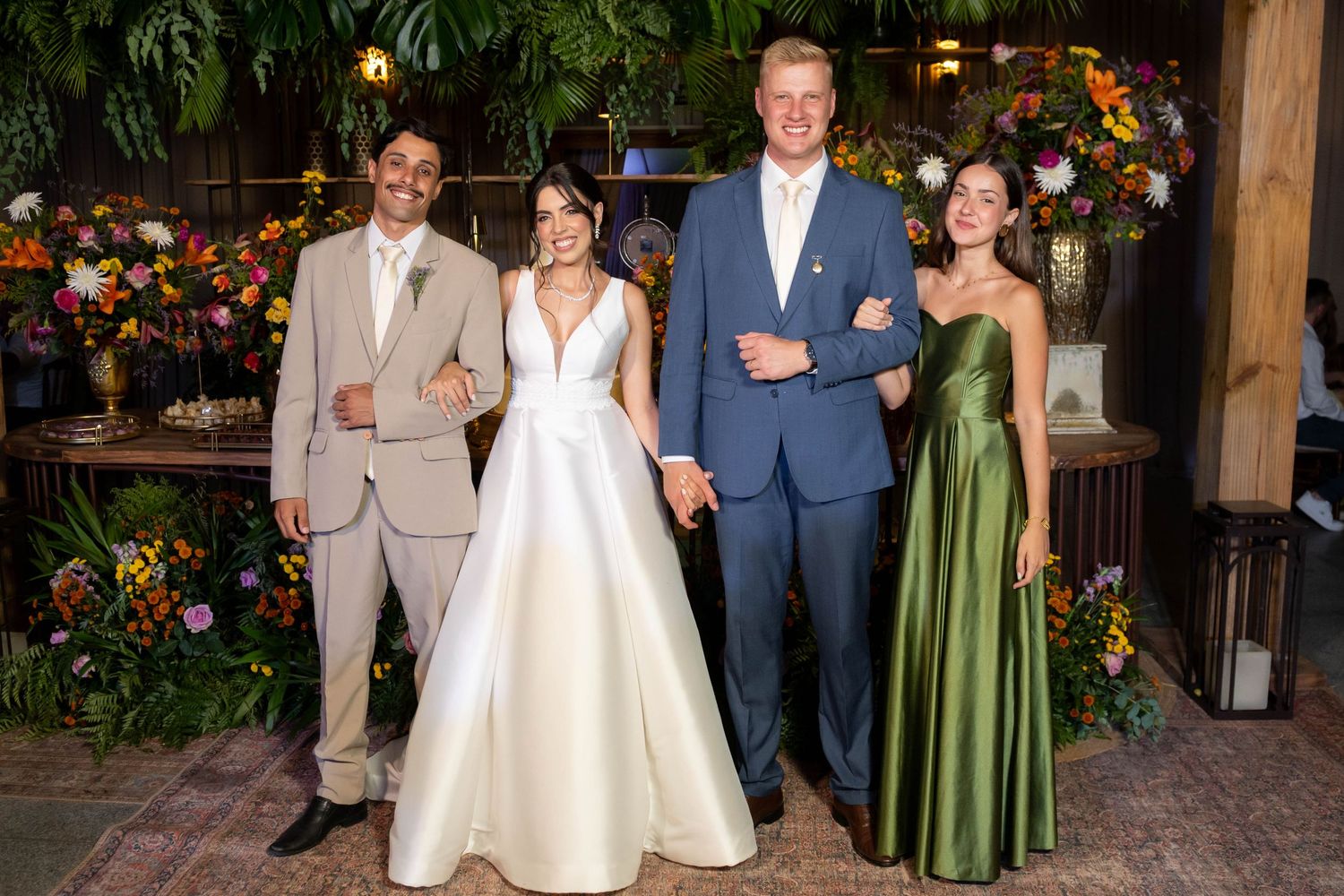 Wedding party of four posing elegantly before a colorful floral backdrop, bride in white gown, guests in formal attire.