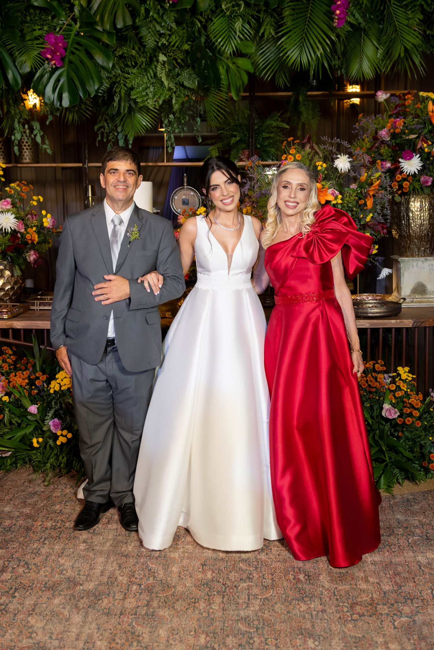 Bride in white gown poses with man in gray suit and woman in red dress at floral wedding reception.