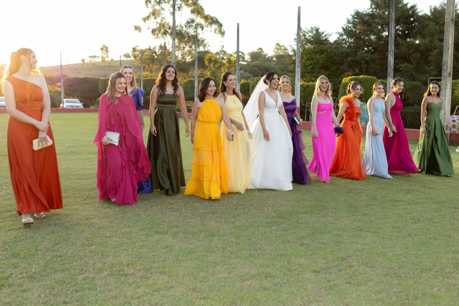 Bride and bridesmaids in rainbow-colored gowns standing on green lawn at outdoor wedding ceremony.