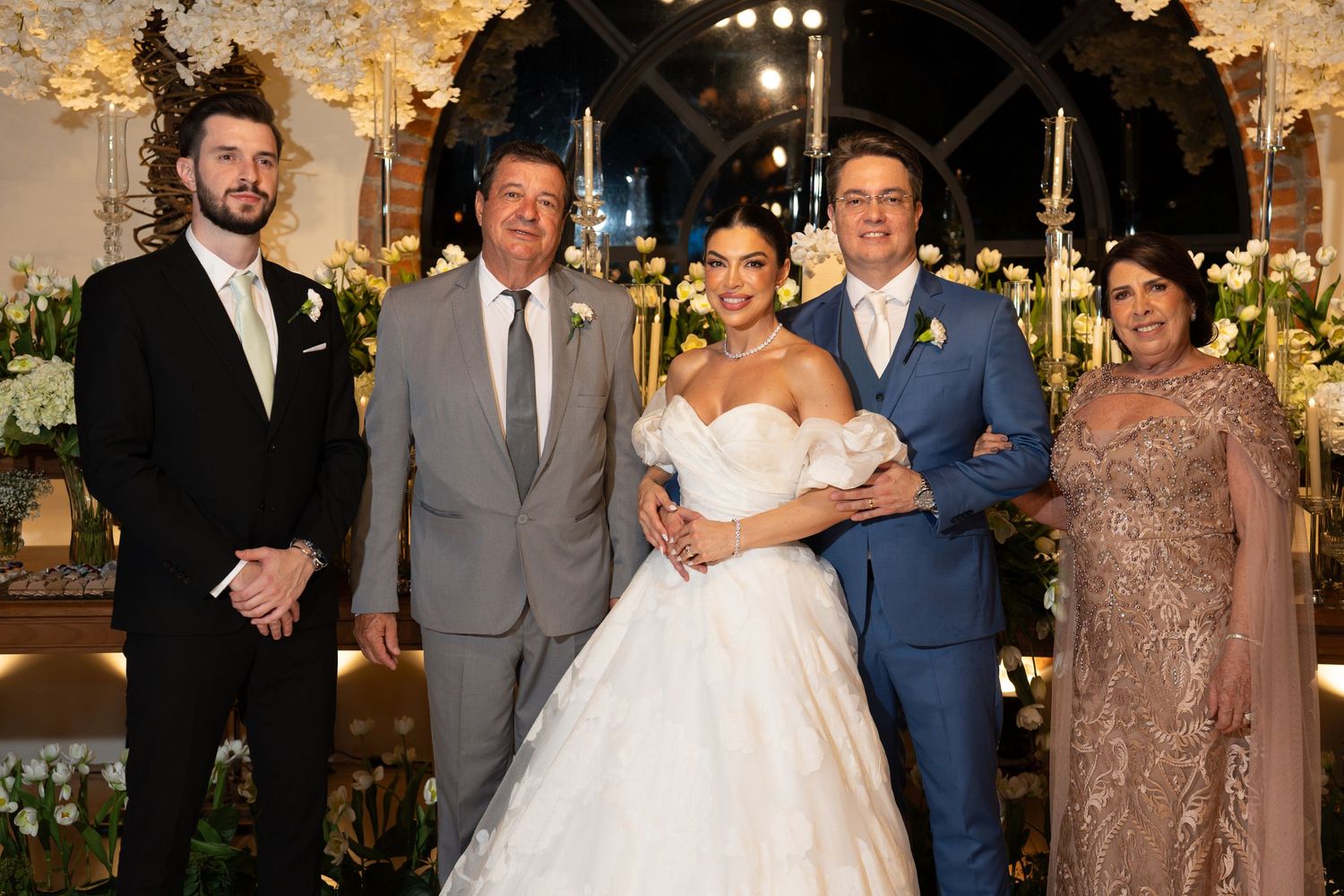 A bride in a white gown poses with groom and family members at an elegant floral wedding ceremony.