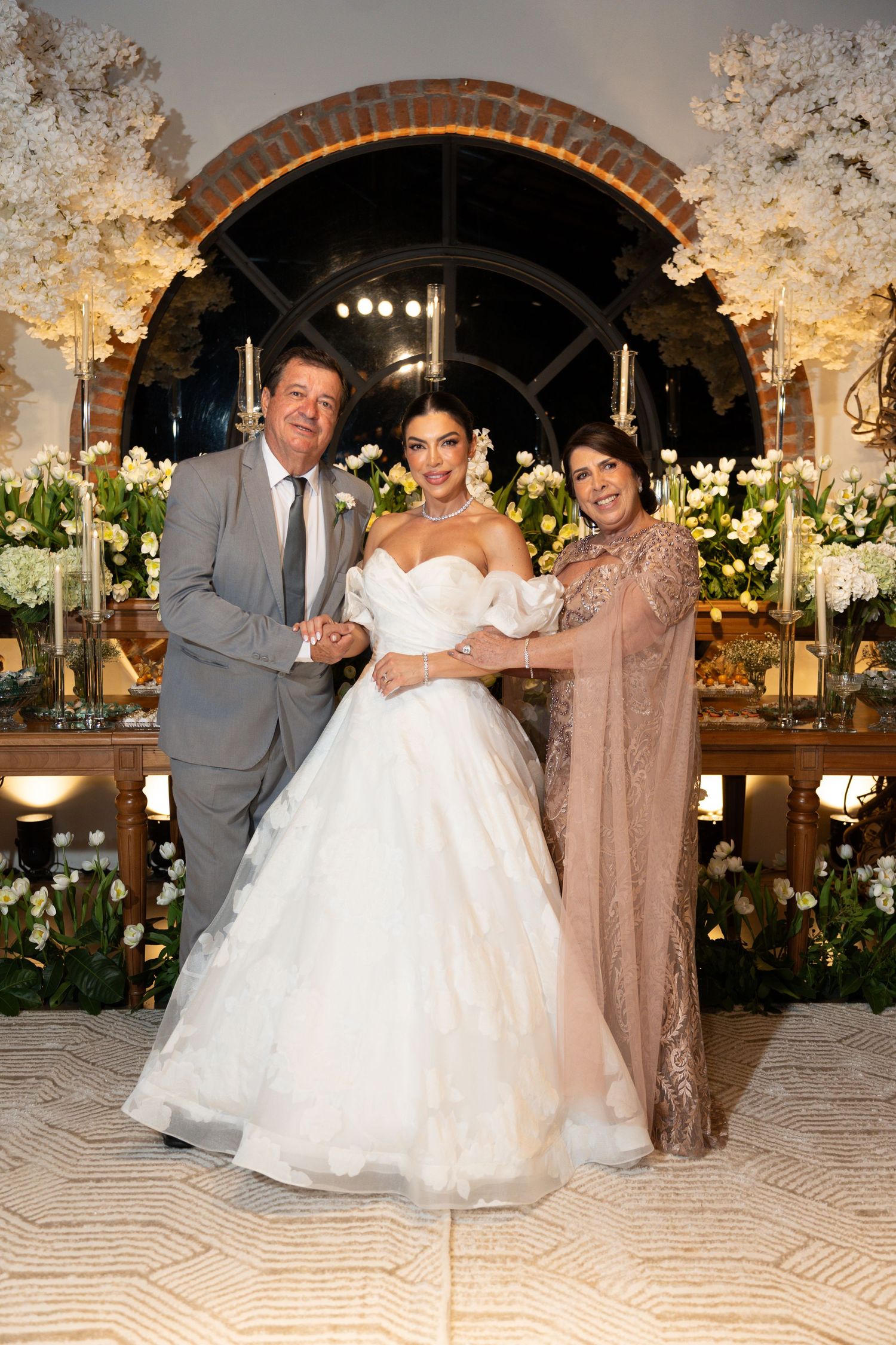 Bride in white ball gown poses with parents at elegant wedding reception with floral arrangements.