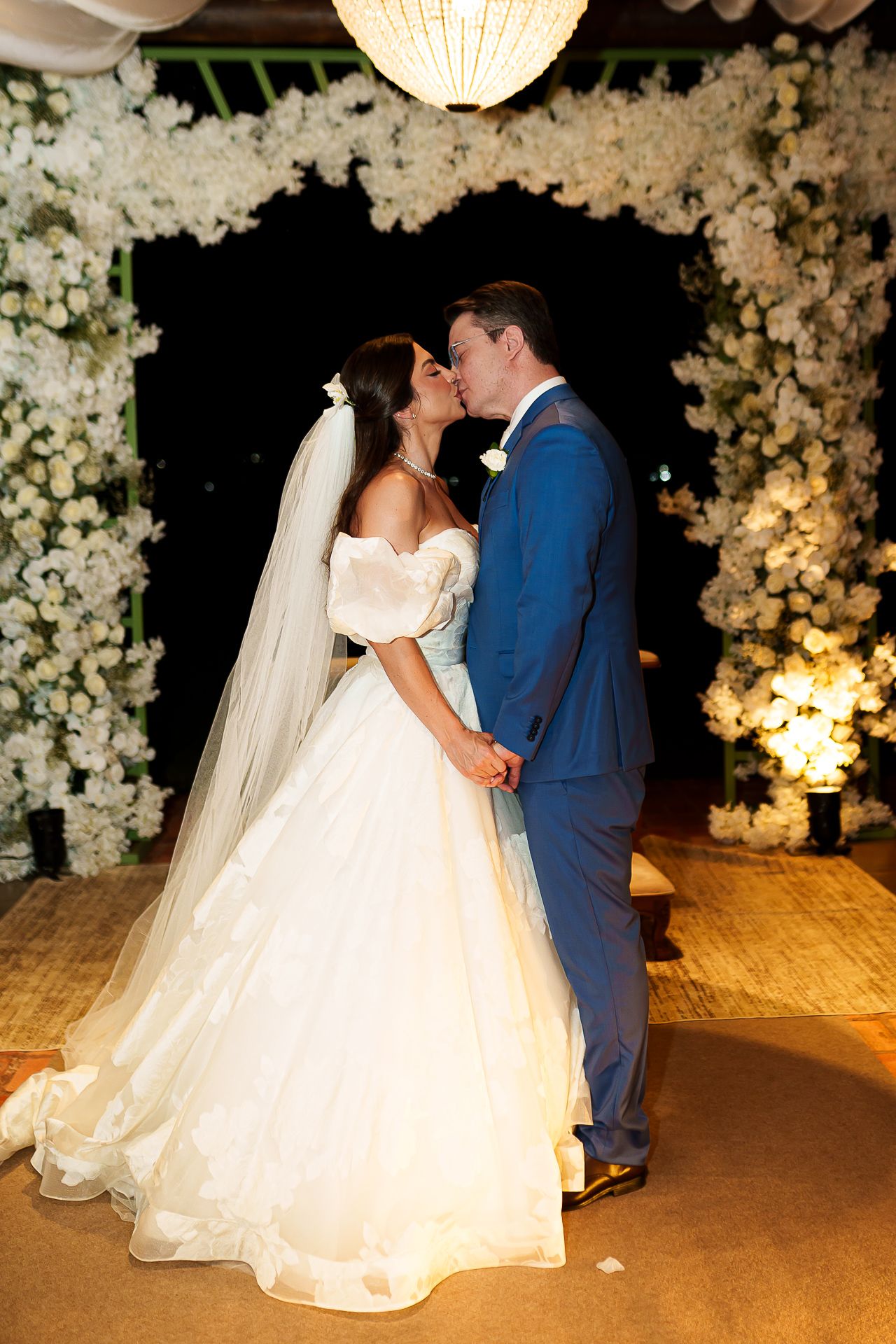 Bride and groom share a first kiss at elegant wedding ceremony surrounded by lush white floral arrangements.