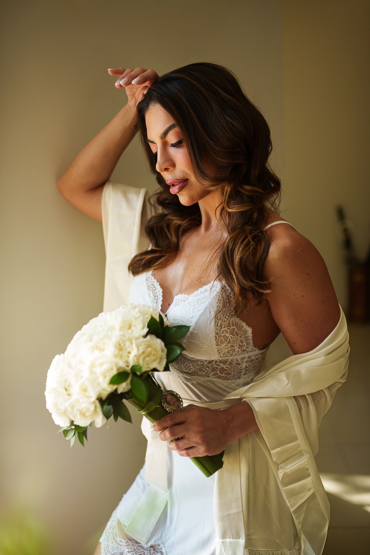 Bride in white lace lingerie and silk robe holds elegant white rose bouquet, posing gracefully in soft natural light.