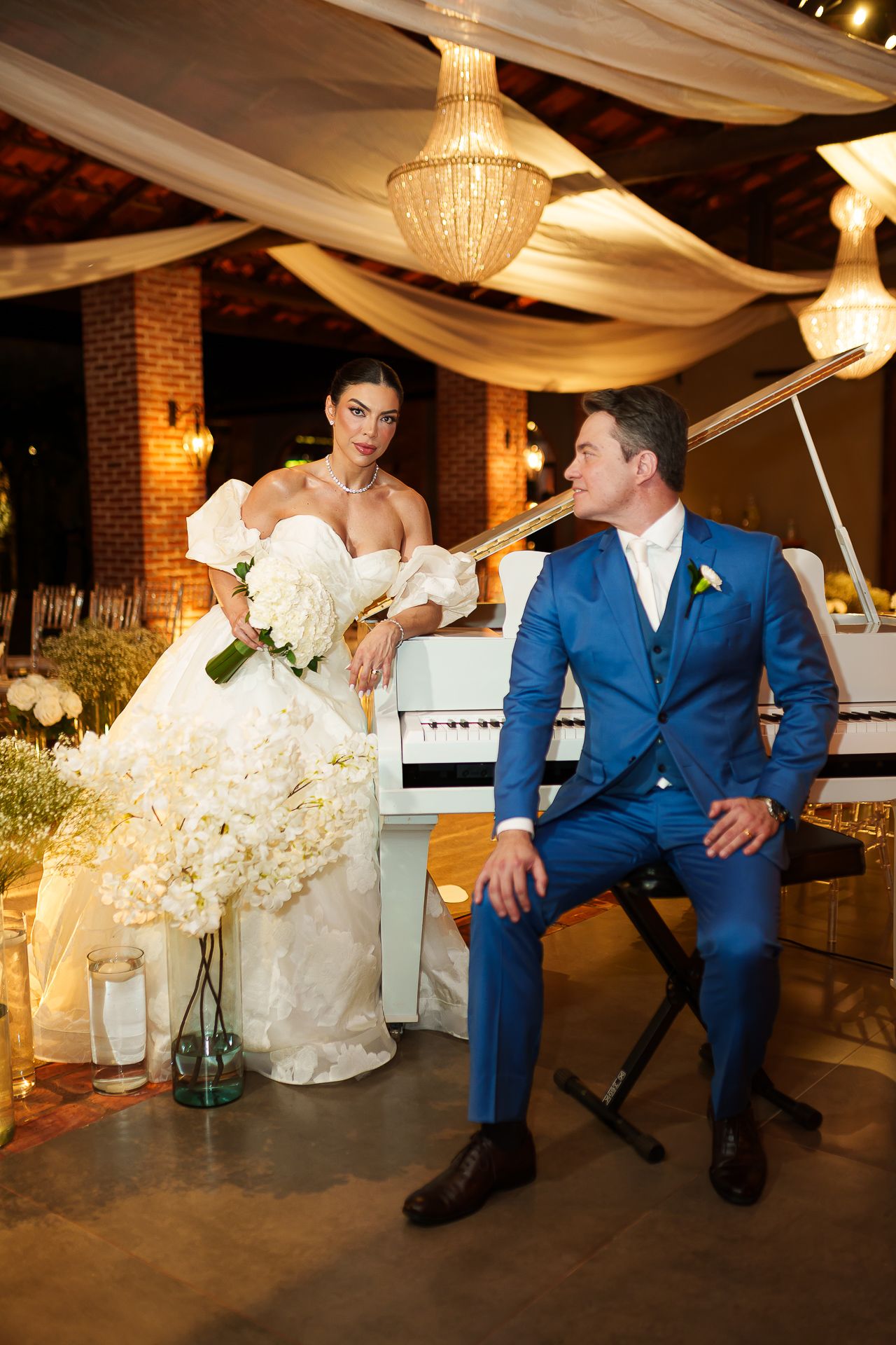 Bride in white gown holds bouquet beside groom in blue suit near white piano under chandelier at elegant wedding venue.