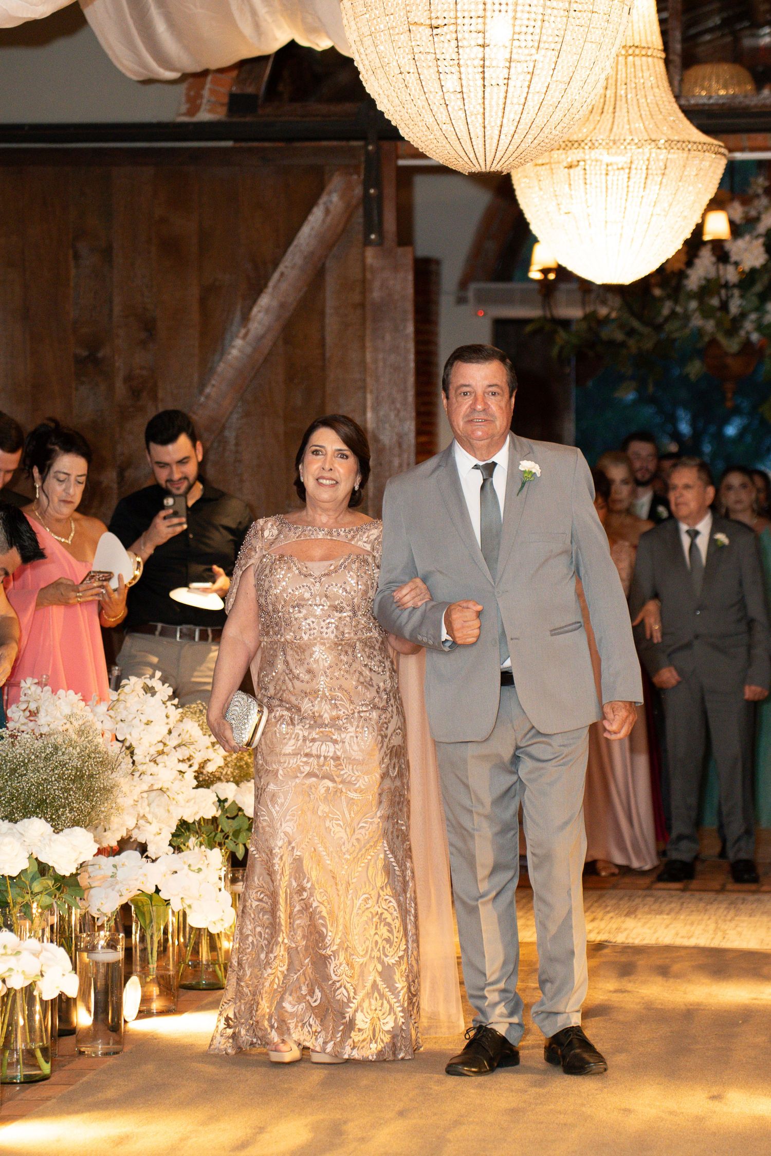 Elegant couple walking down wedding aisle, woman in gold sequin gown, man in gray suit with boutonniere, chandelier backdrop.