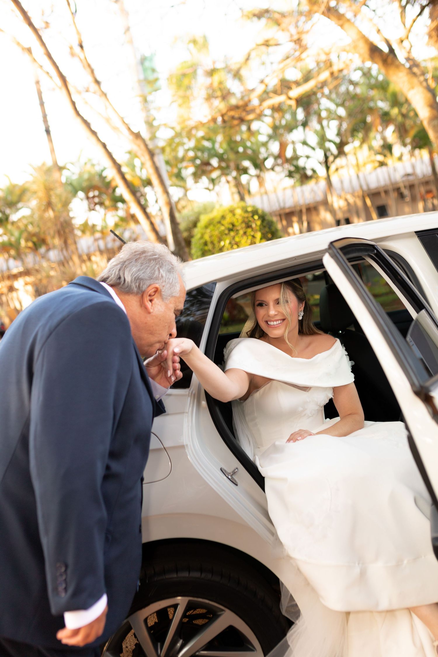 Elegant older bride in off-shoulder white gown exits a white car as a gentleman kisses her hand.