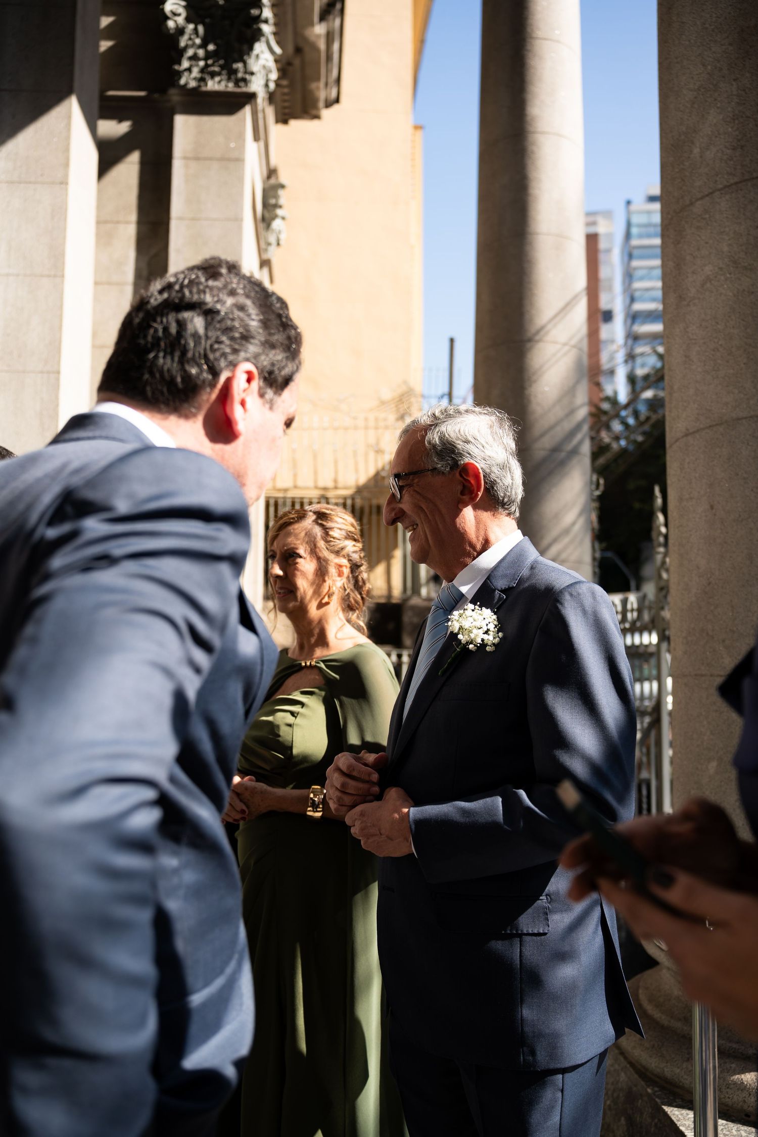 Elegantly dressed guests in formal attire converse outside a church at a sunny wedding celebration.