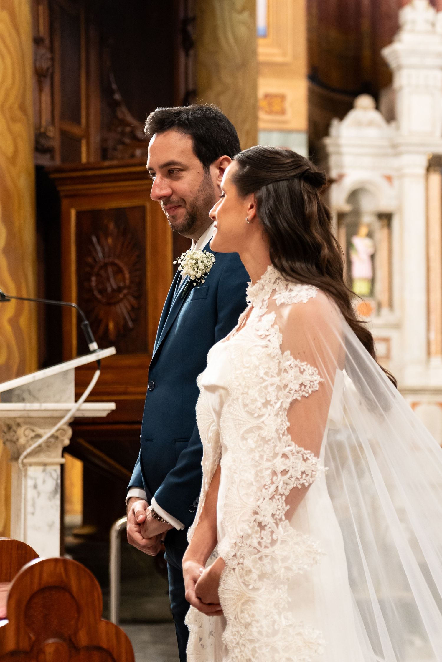 Bride and groom holding hands at church altar, bride in lace gown with veil, groom in navy suit with boutonniere.