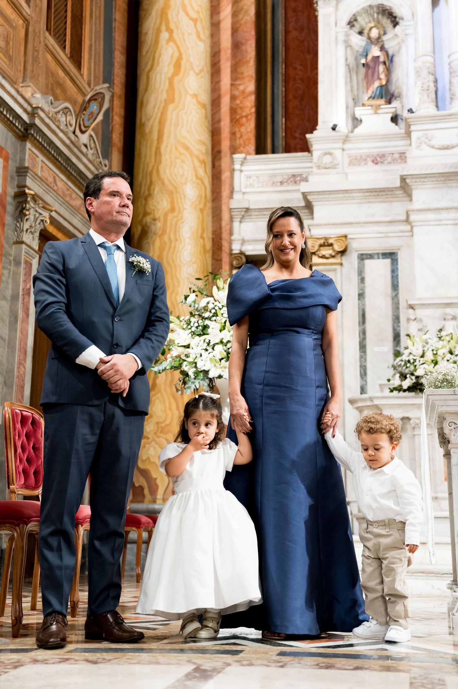Elegant family in formal attire poses inside ornate marble church, woman in navy gown with two young children.