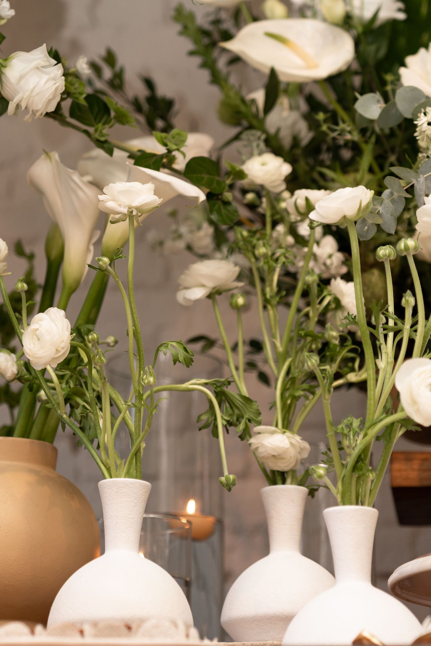 White ranunculus and calla lilies in matte white vases with a glowing candle on a decorated table.