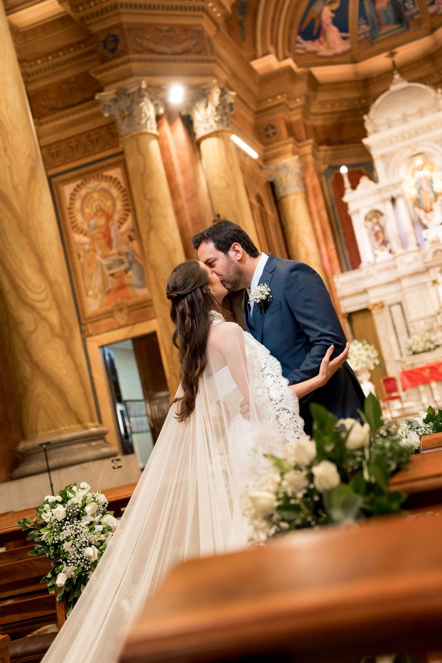 A bride and groom share their first kiss at the altar inside an ornate church decorated with white floral arrangements.