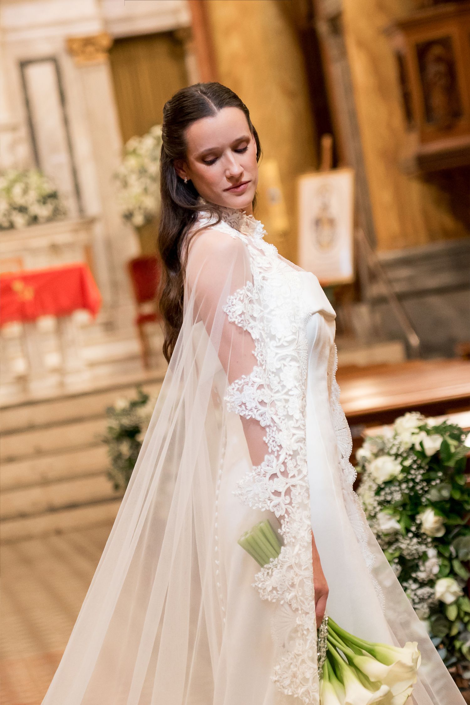 Elegant bride in lace wedding gown with sheer cape veil holding white bouquet inside ornate church.