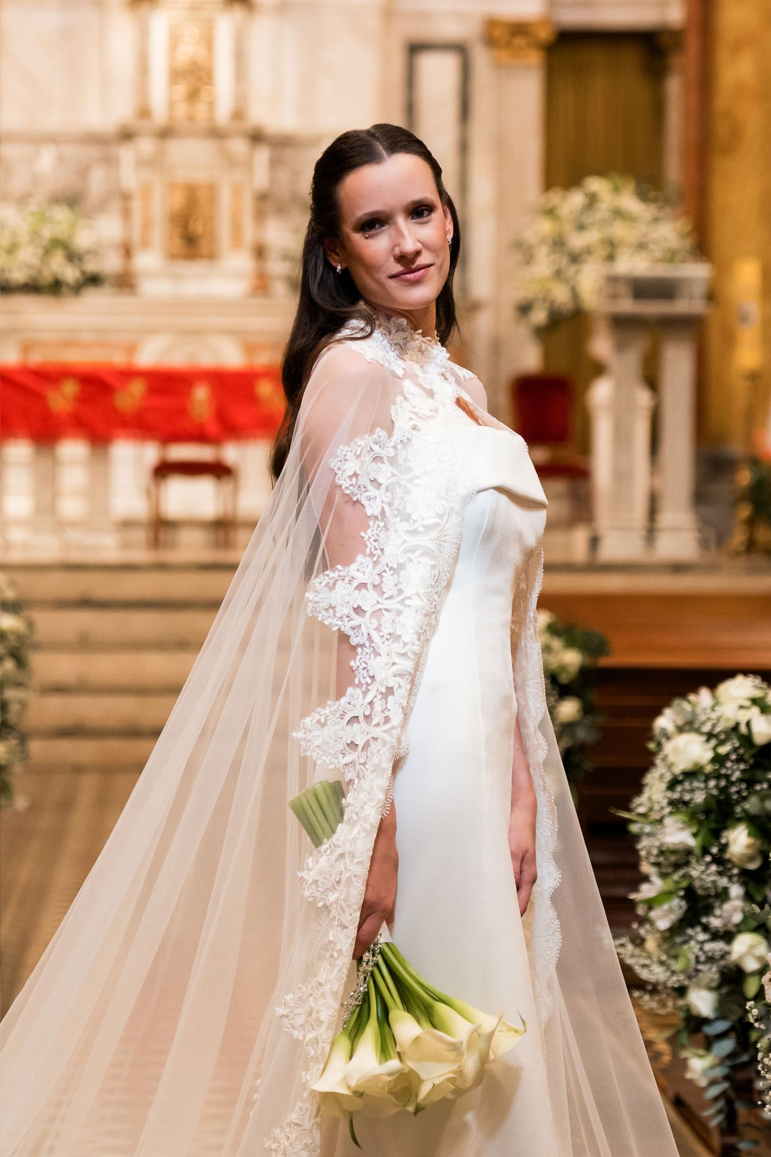 Elegant bride in white gown with lace veil holds calla lily bouquet inside ornate church altar.