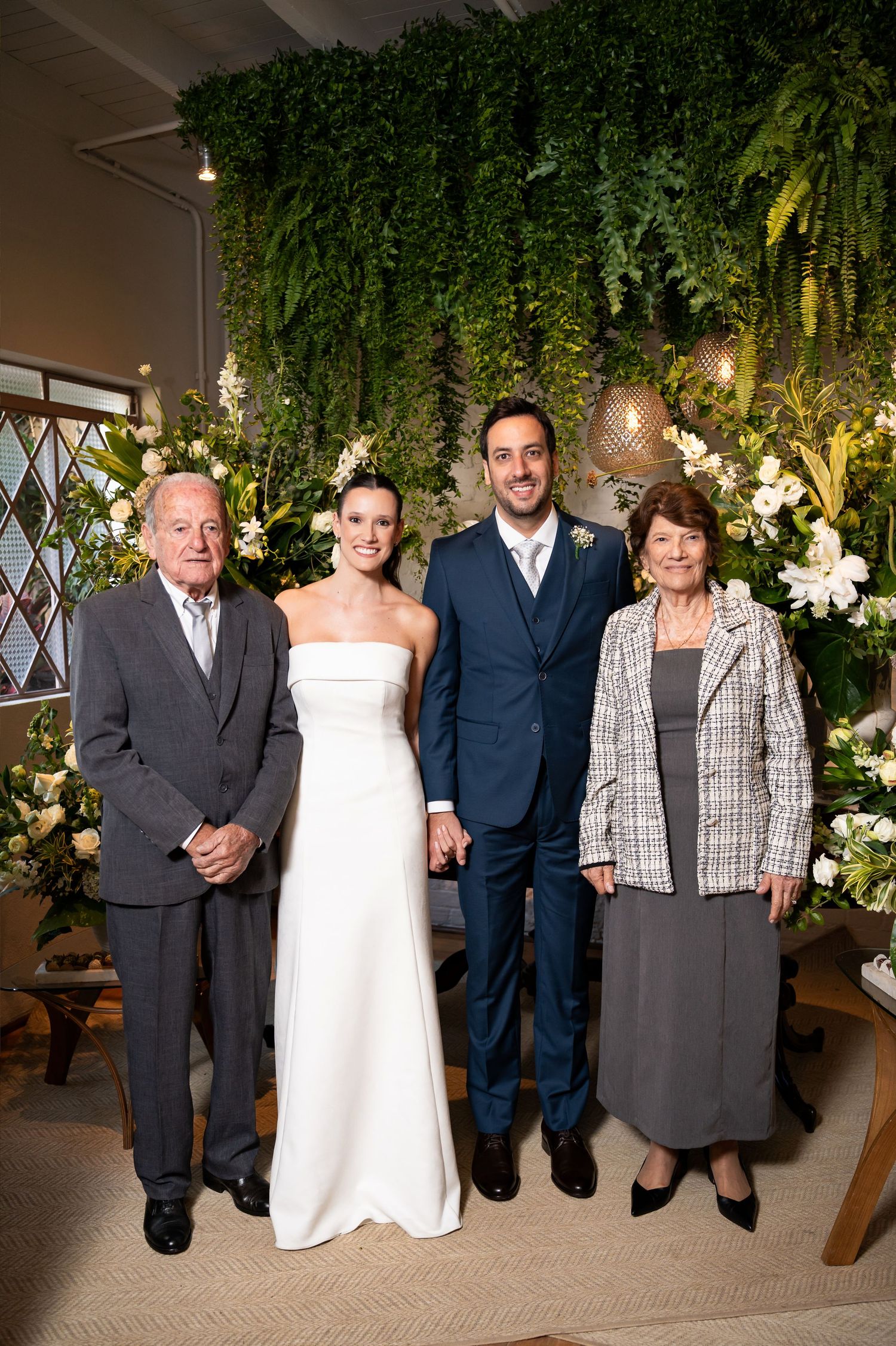 Bride in white gown and groom in navy suit pose with elderly couple at floral wedding ceremony backdrop.