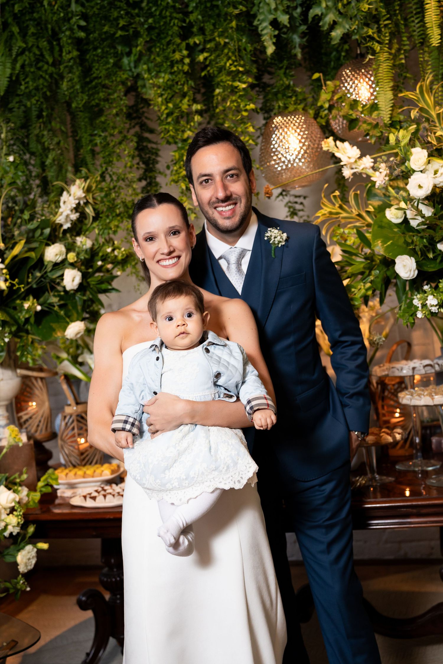 Happy couple in wedding attire smiling and holding their baby at an elegant floral wedding reception.