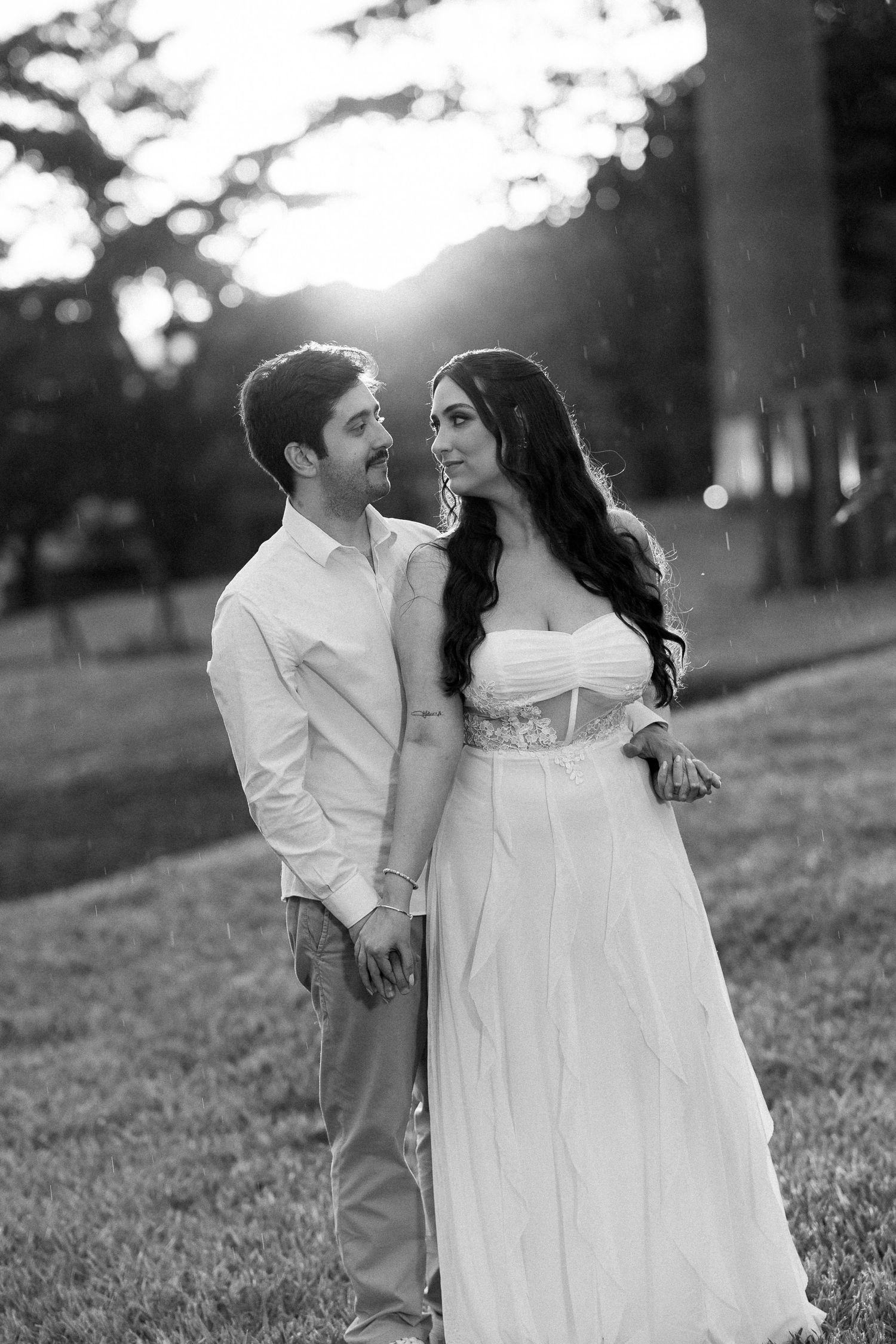 Black and white photo of a couple in wedding attire gazing at each other in a sunlit outdoor setting.