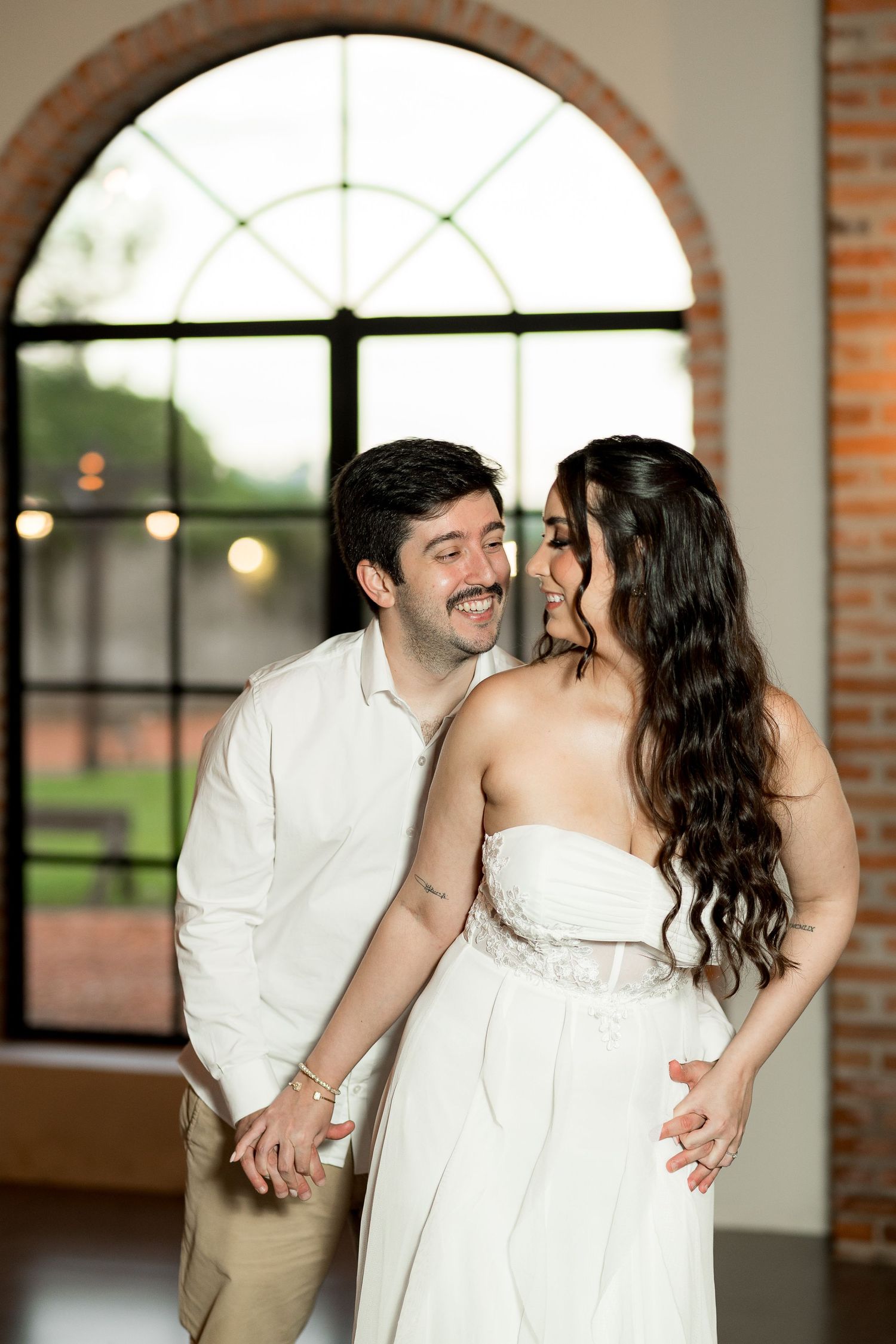 A smiling couple in white attire holds hands near an arched window in a brick venue.