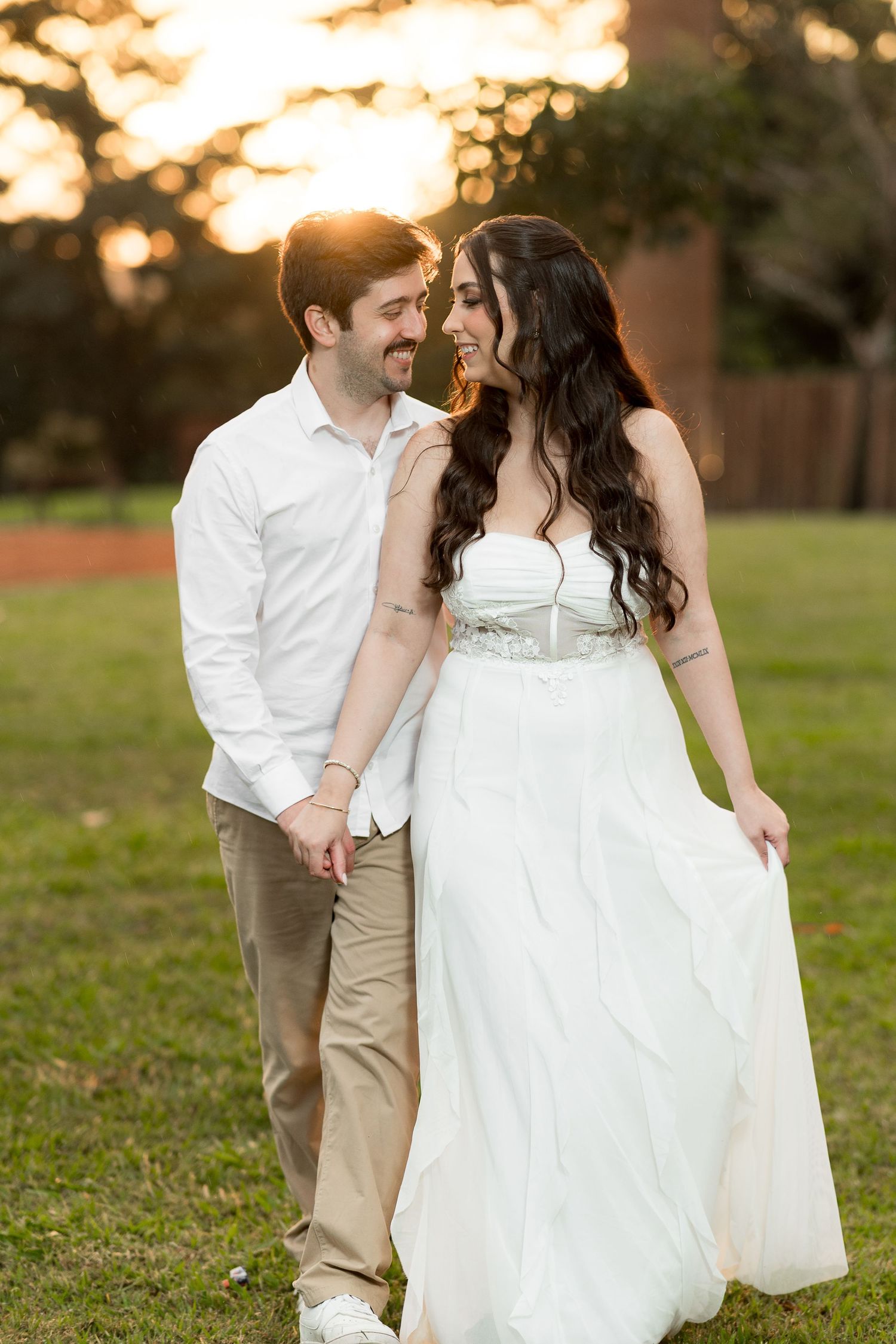 Couple in love walking on grass at sunset, woman in white wedding dress, man in white shirt and khaki pants.