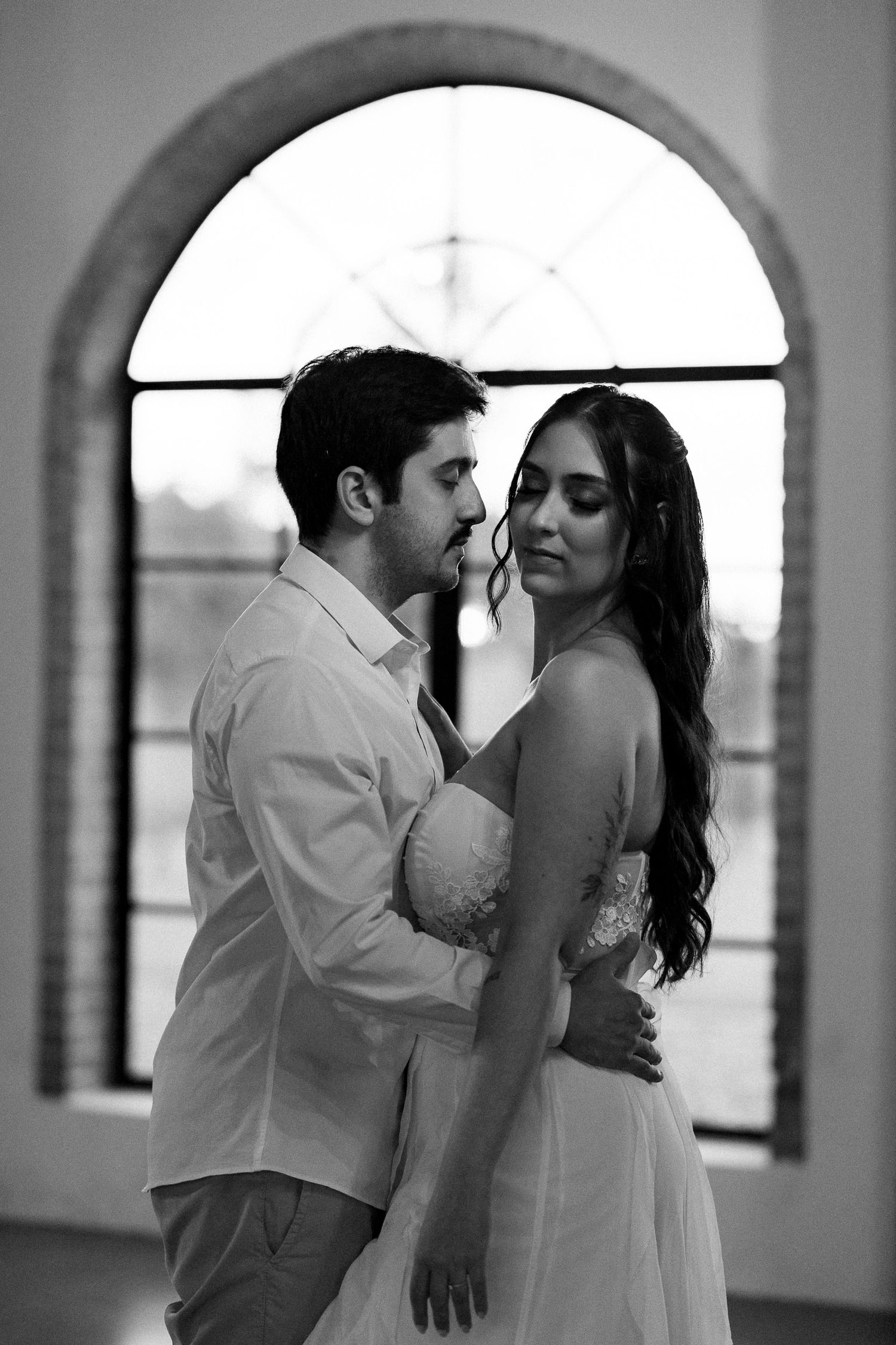 Black and white photo of a couple embracing romantically in front of an arched window in elegant attire.