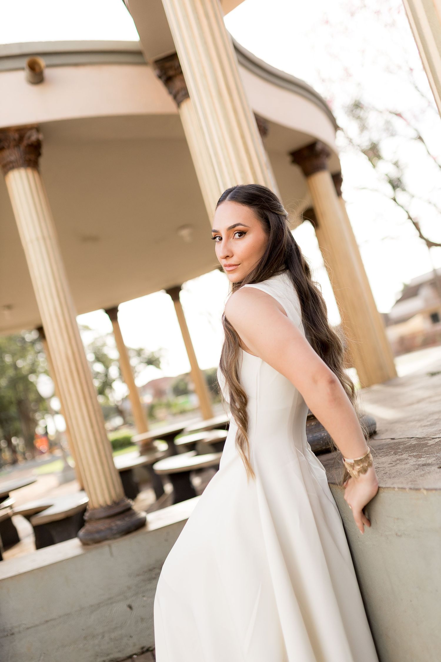 Young woman in white dress posing by classical columns at an elegant outdoor pavilion.