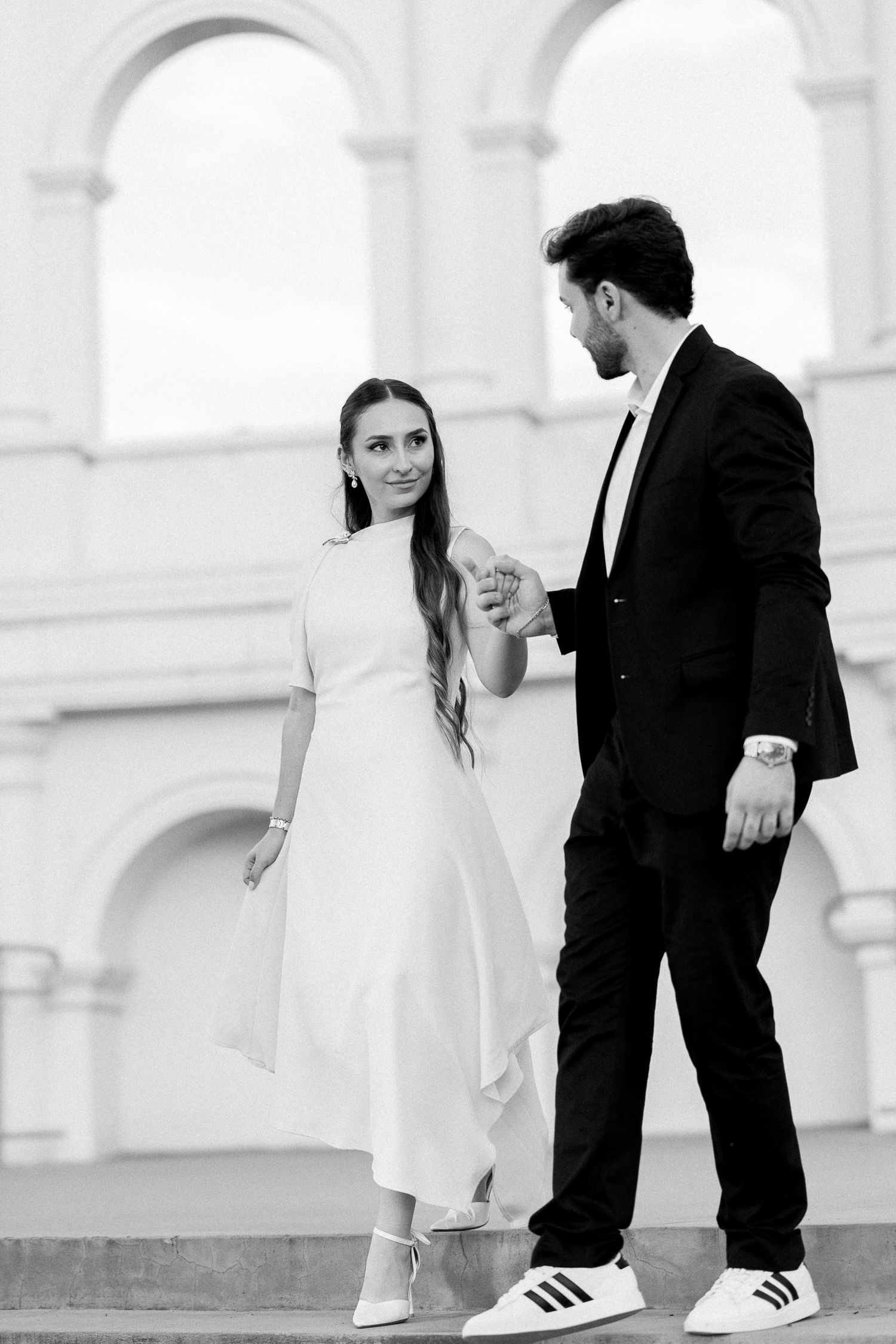 Black and white photo of an elegant couple holding hands on steps, woman in white dress, man in dark suit.