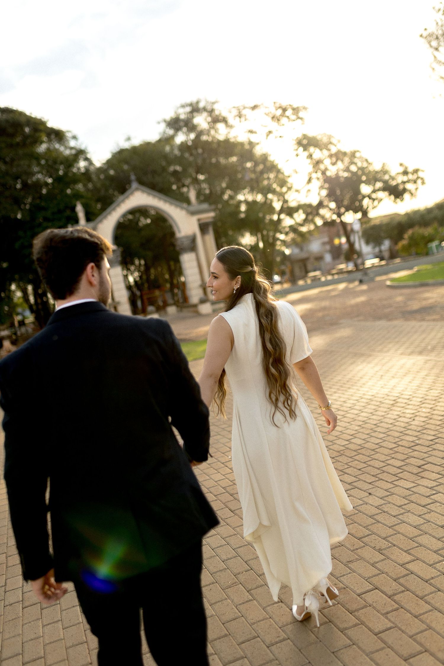 Bride in white dress and groom in black suit walk hand in hand on brick path toward arch at golden hour.