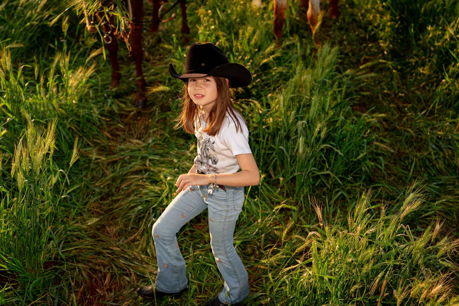 Young girl in black cowboy hat, white tee, and flare jeans poses playfully in lush green field at golden hour.