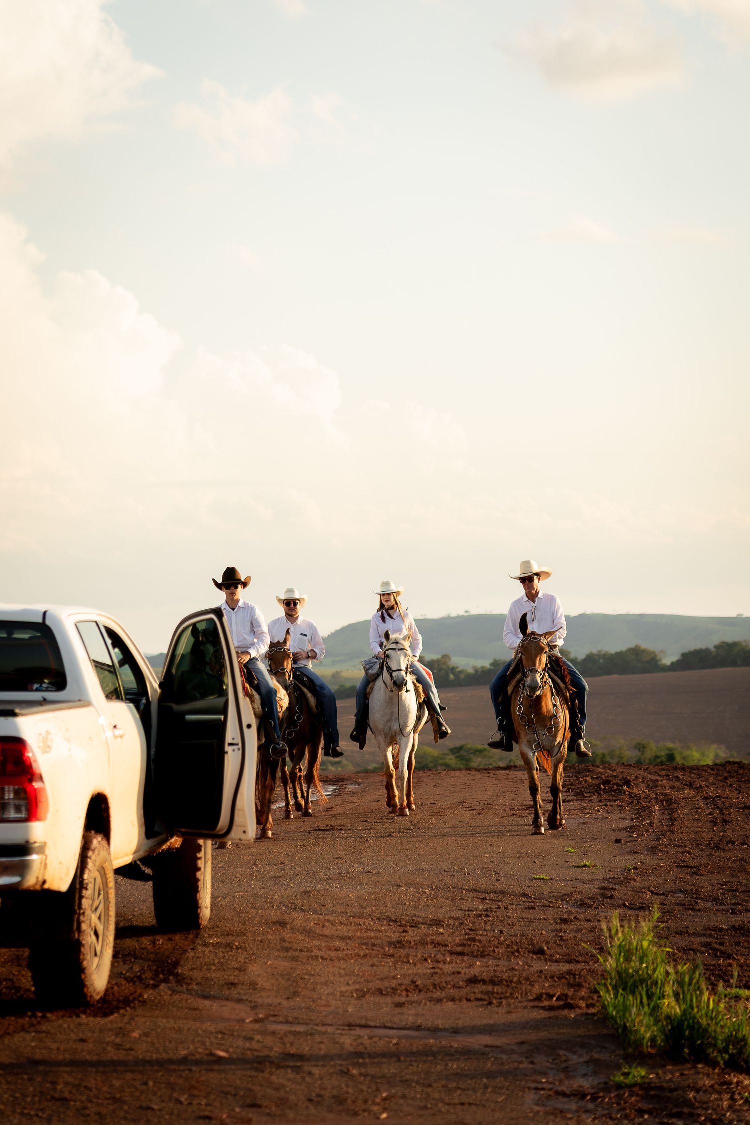Four cowboys on horseback ride along a dirt road past a white pickup truck in a rural landscape.
