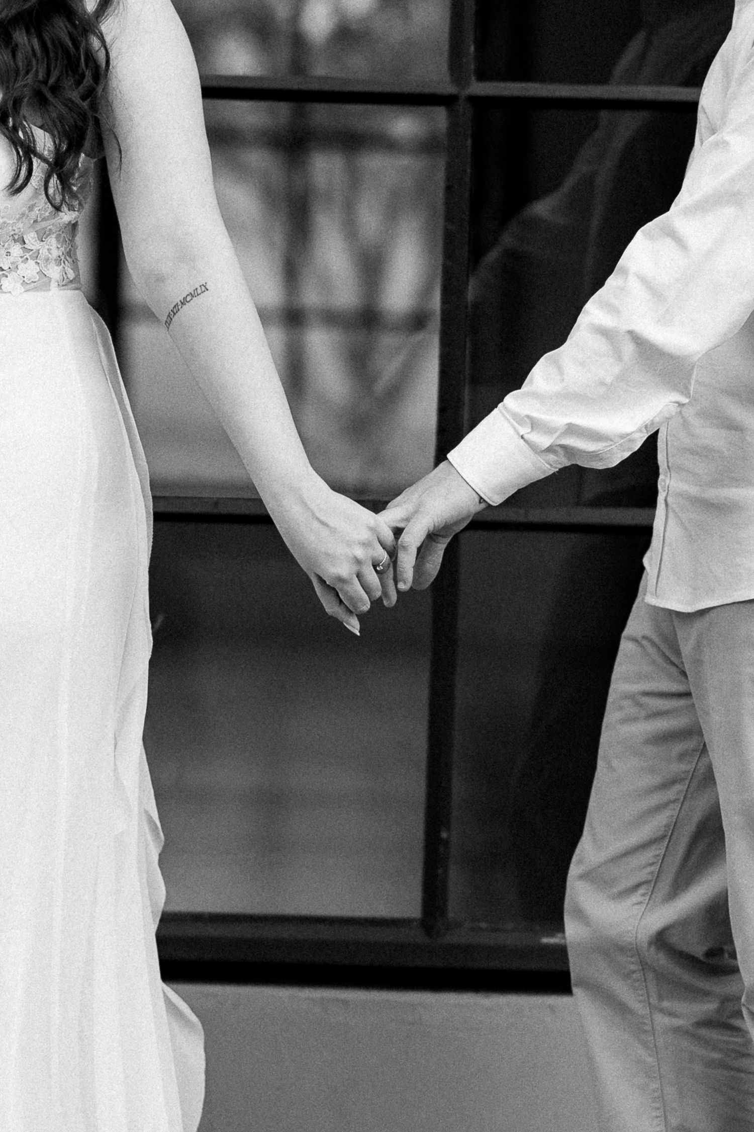 Black and white photo of a bride and groom holding hands in front of a window on their wedding day.
