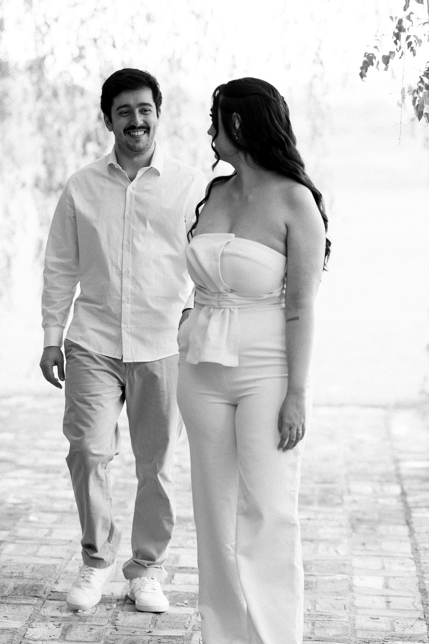 Black and white photo of a smiling couple walking together outdoors near a waterfront, dressed in casual white outfits.