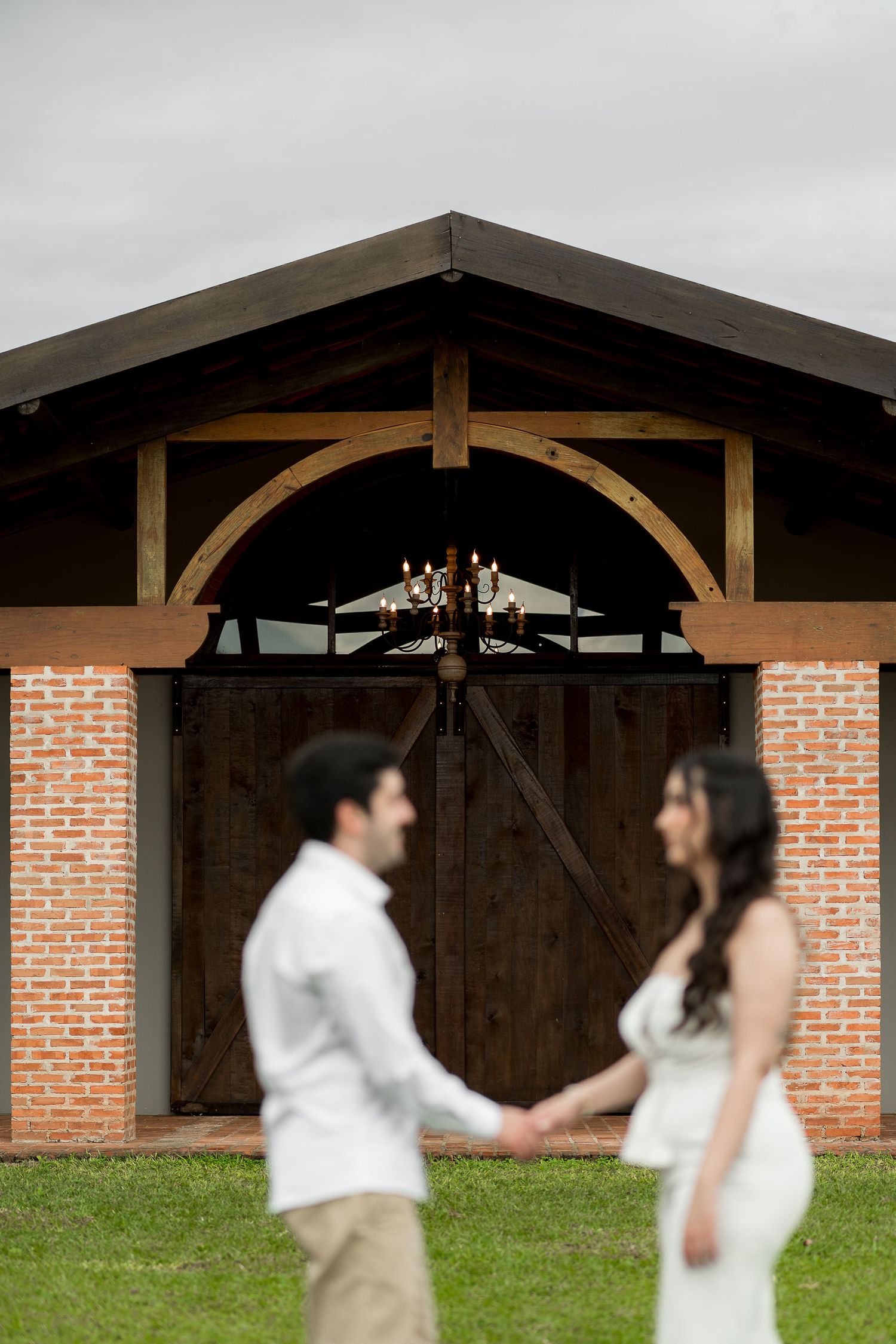 Couple holds hands in front of rustic brick barn with wooden doors and chandelier at wedding venue.