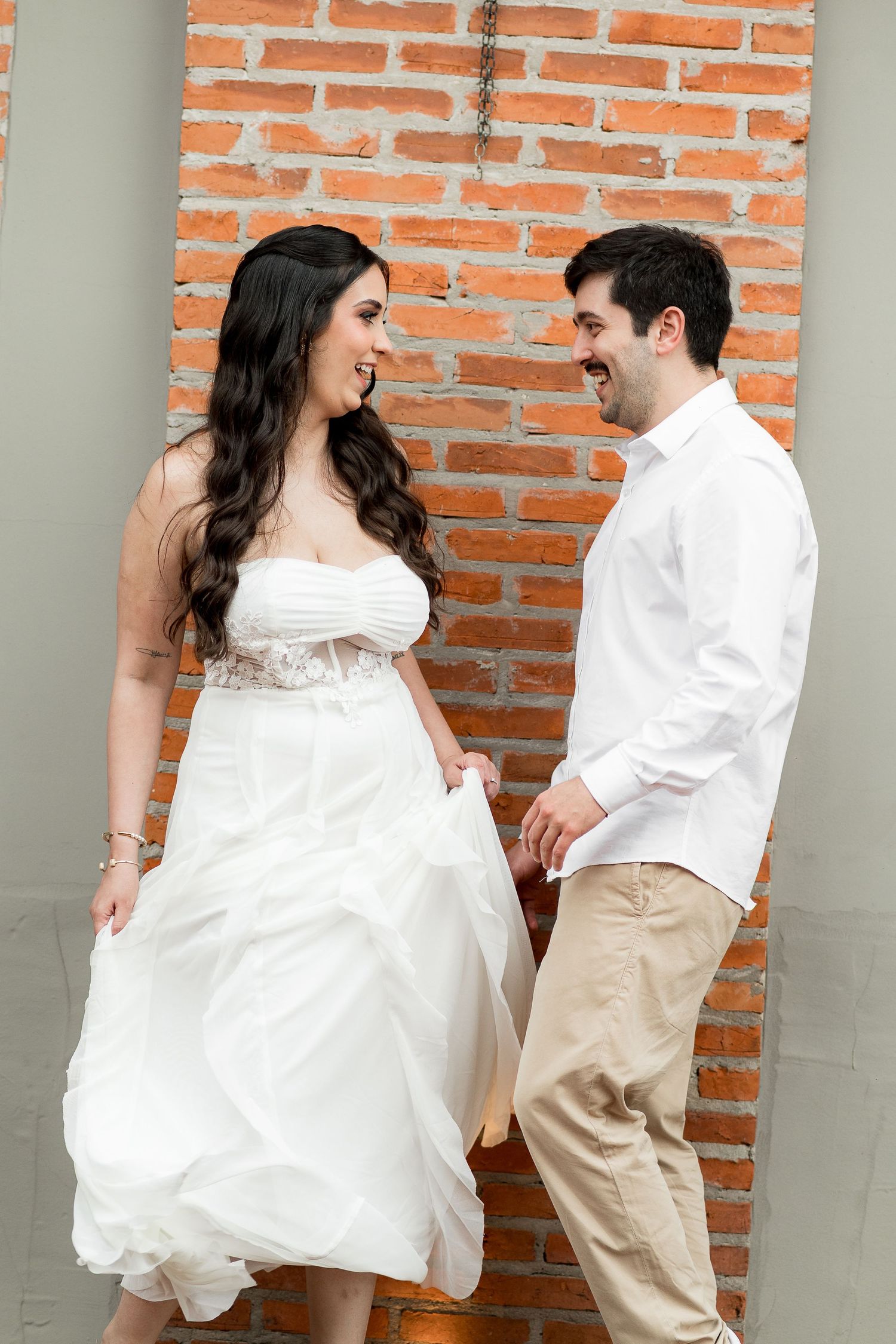 Smiling couple in wedding attire holding hands in front of a red brick wall, bride in white gown and groom in white shirt.