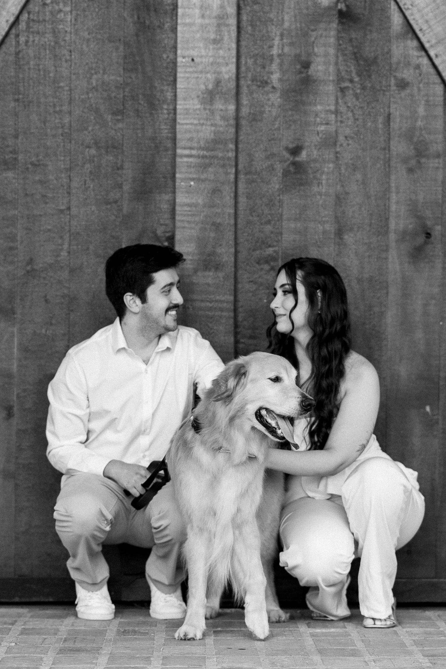 Black and white photo of a smiling couple in white outfits crouching beside a fluffy dog in front of a wooden wall.