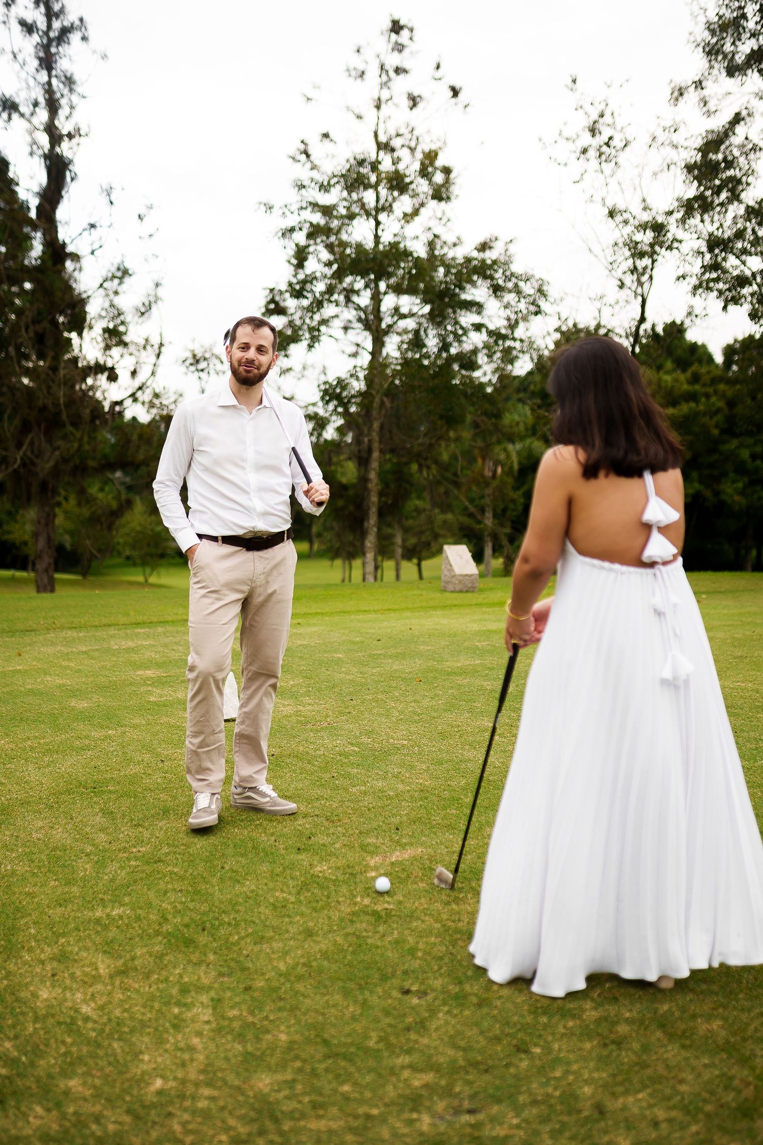 Bride in white backless gown playing golf with groom on lush green course.