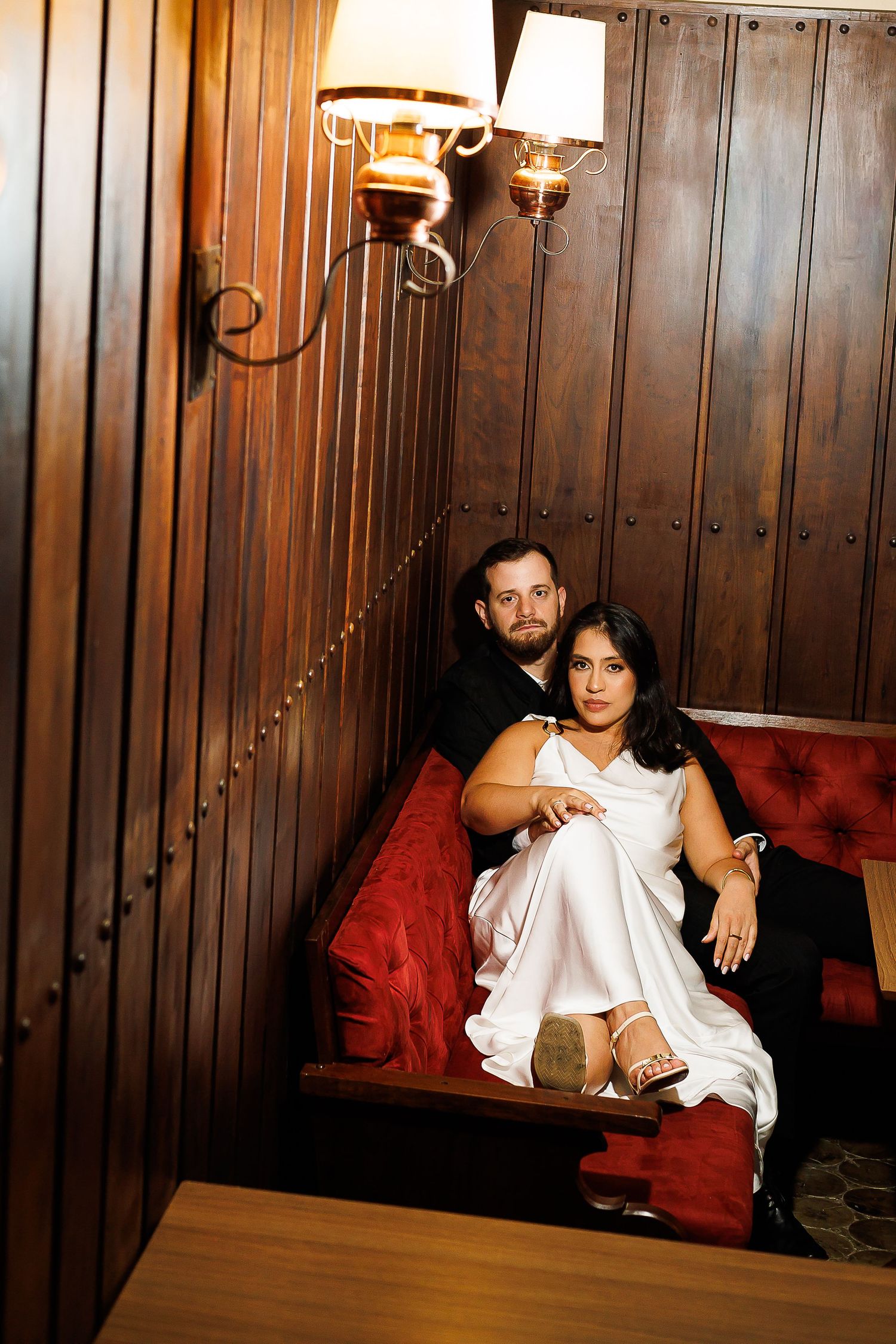 Couple in elegant attire sitting on red velvet booth in a warmly lit wood-paneled restaurant lounge.