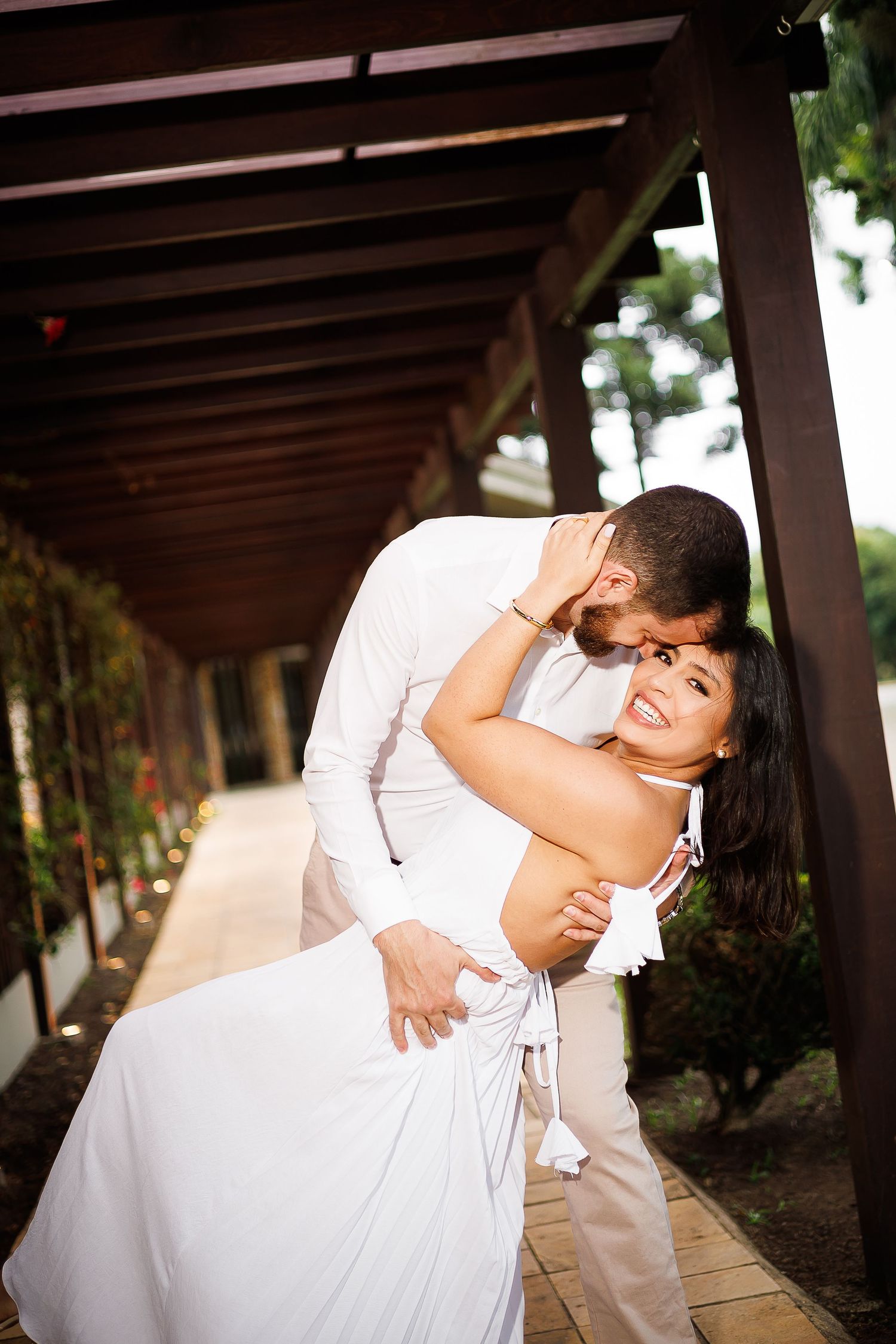 Couple in white outfits sharing a romantic embrace under a wooden pergola, woman smiling joyfully.