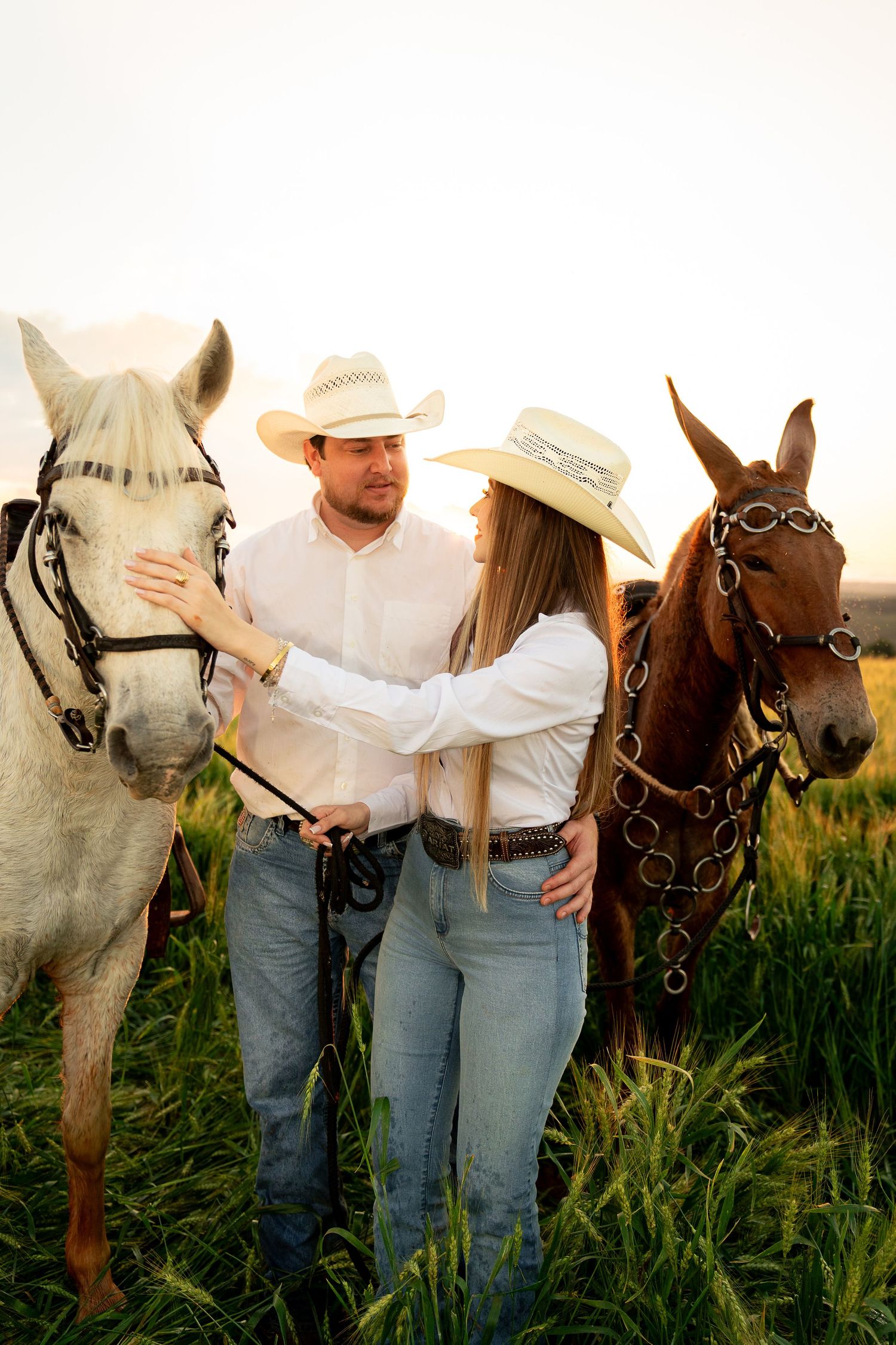A couple in cowboy hats and jeans stand together in a field with a white and brown horse at golden hour.